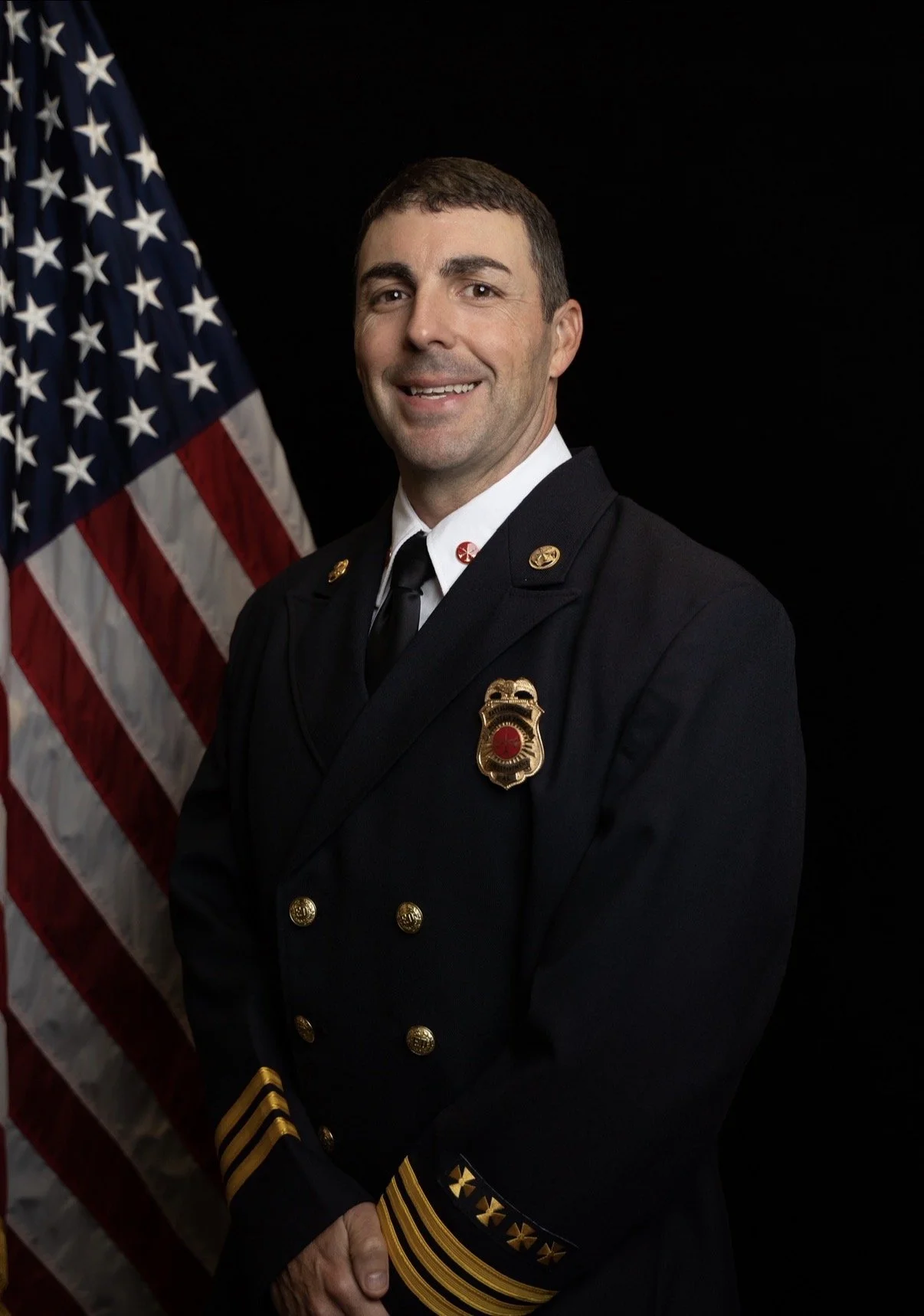 A male firefighter in uniform standing in front of an American flag, smiling on a black background.