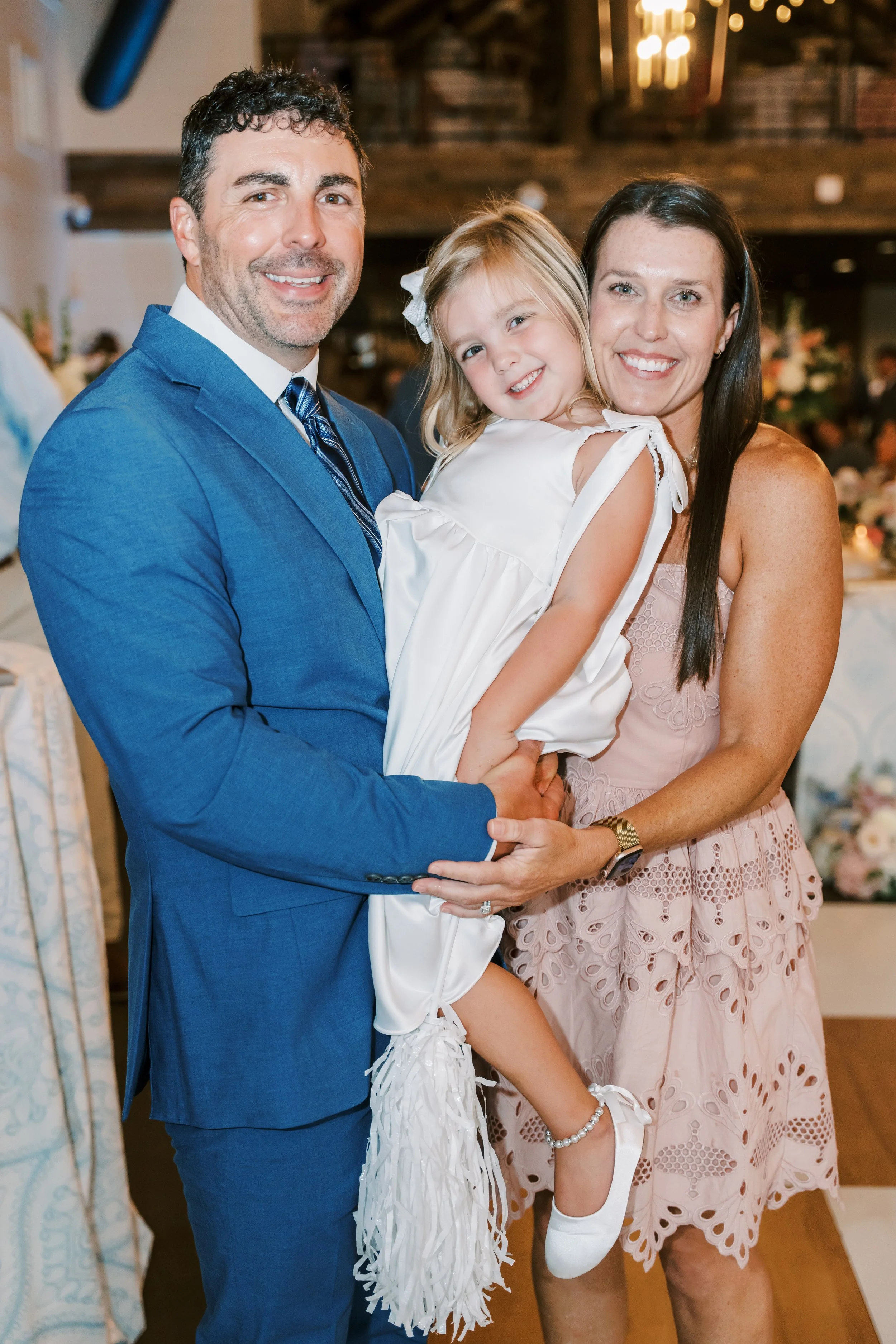 A happy family at a celebration, featuring a man in a blue suit, a woman in a pink dress, and a young girl in a white dress with ruffles. They are smiling and posing closely together in a warmly lit venue with blurred background decorations.