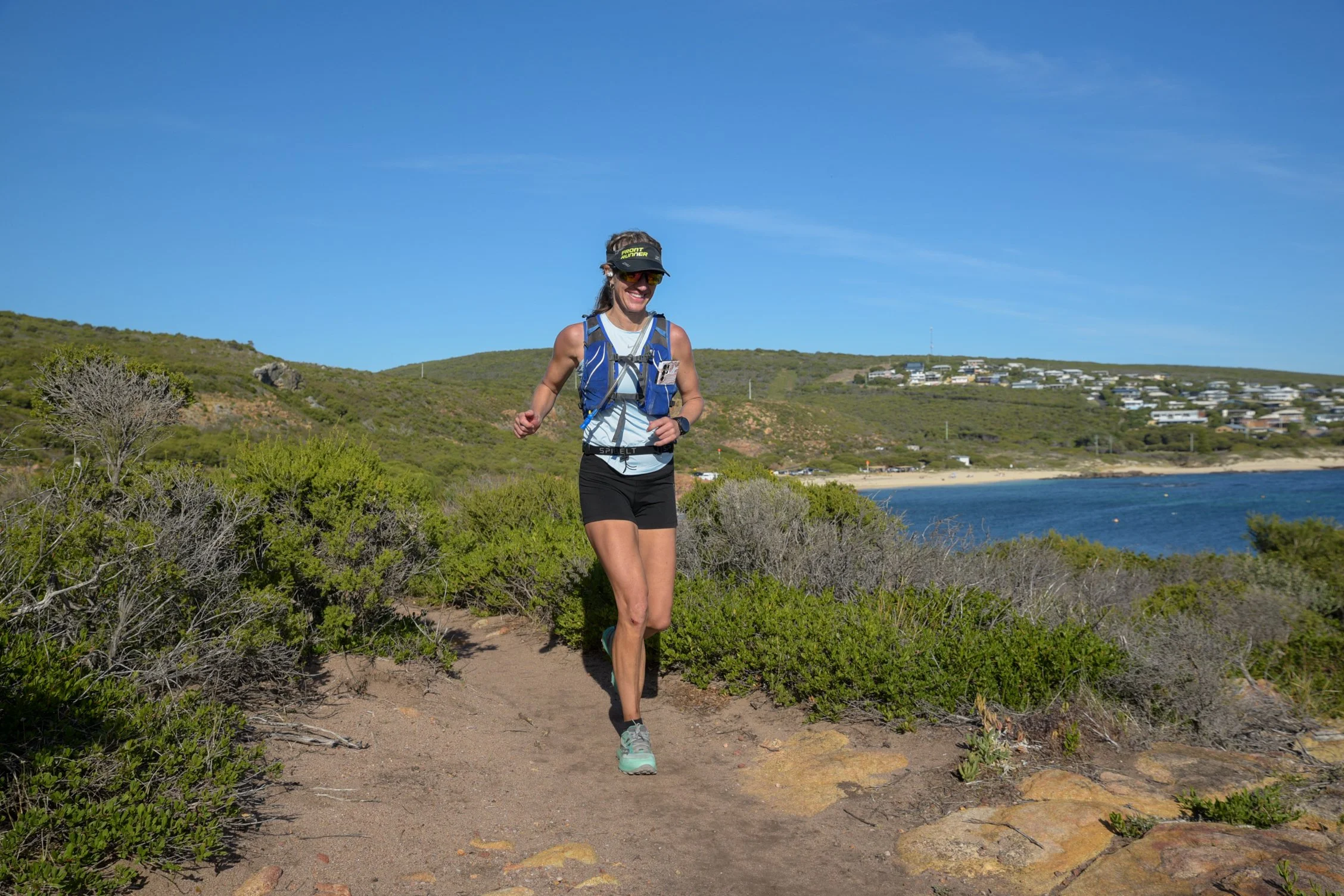 A woman running on a trail along a coastal landscape under a clear blue sky, wearing athletic gear and a hiking backpack.