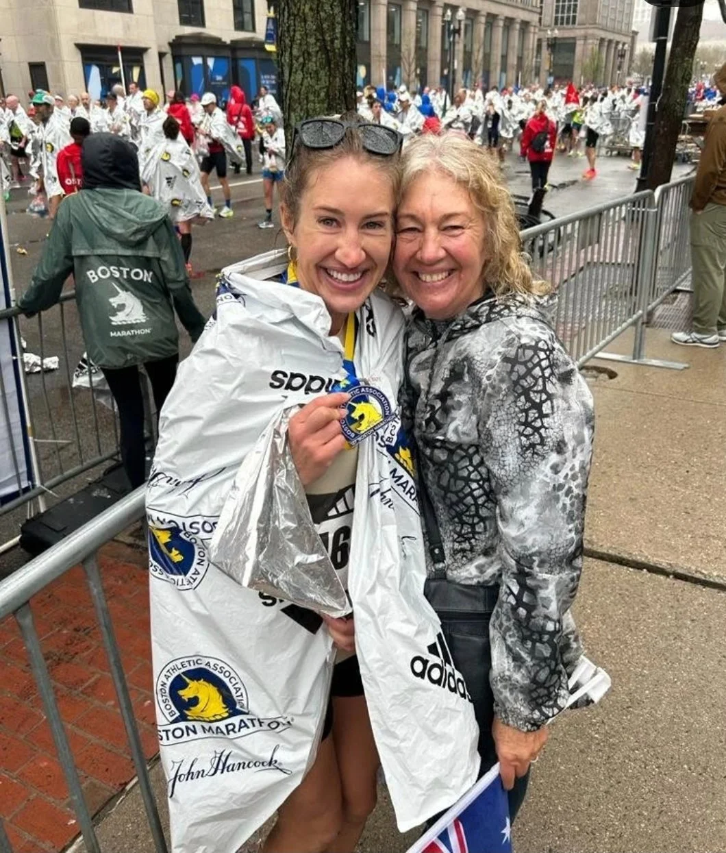 Two smiling women, one wearing a Boston Marathon poncho and medal, celebrating after a race, with a large crowd in the background.