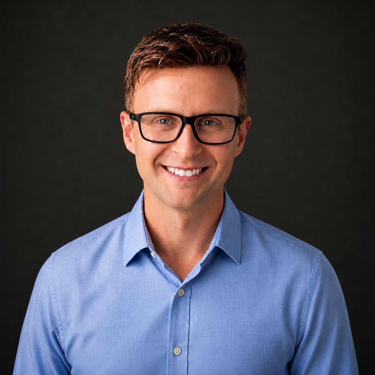A smiling man wearing glasses and a light blue button-up shirt, standing against a dark background.