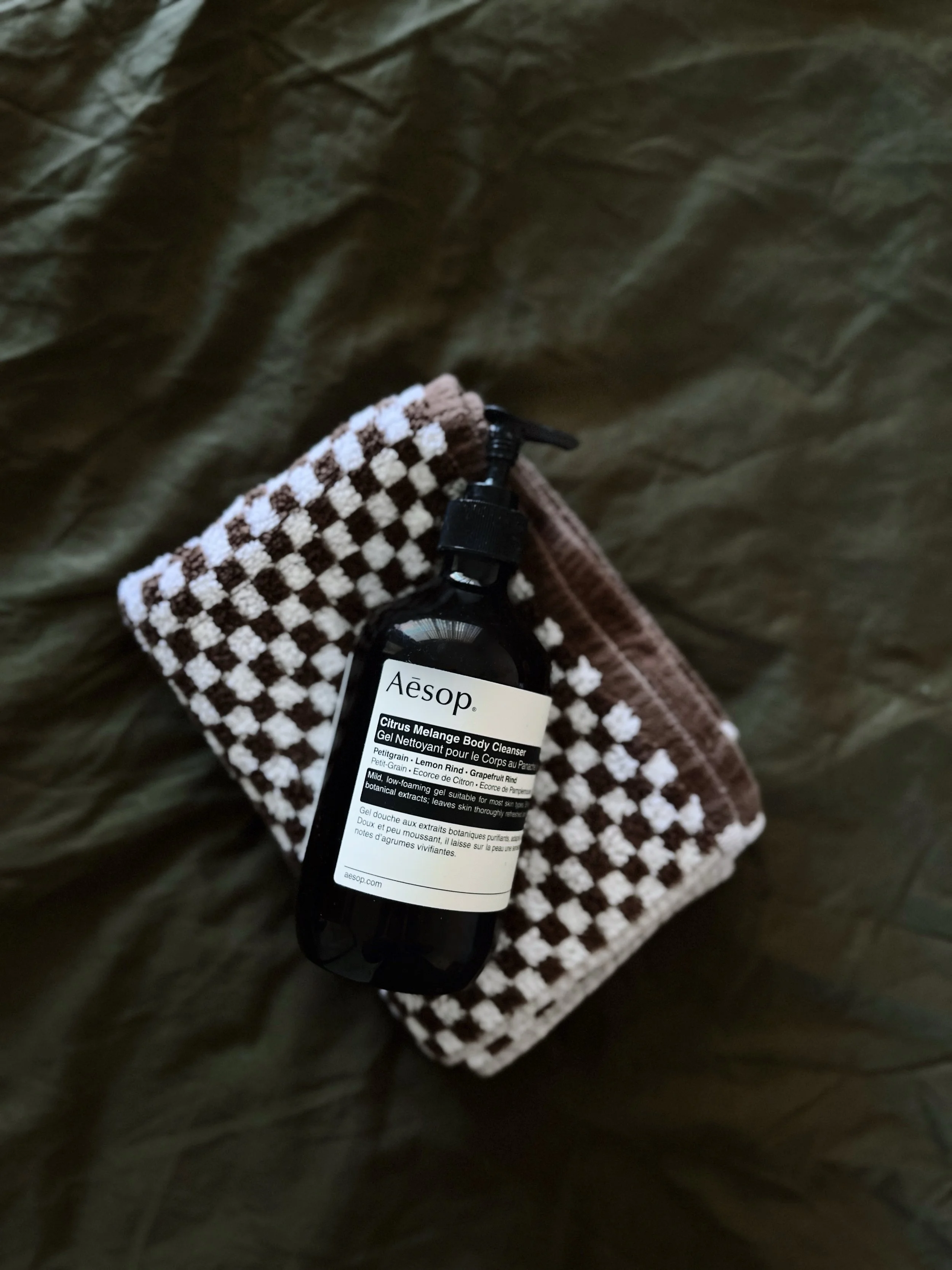 A black bottle of Aesop Citrus Melange Body Cleanser resting on a brown and white checkered cloth, with a background of dark, crinkled fabric.