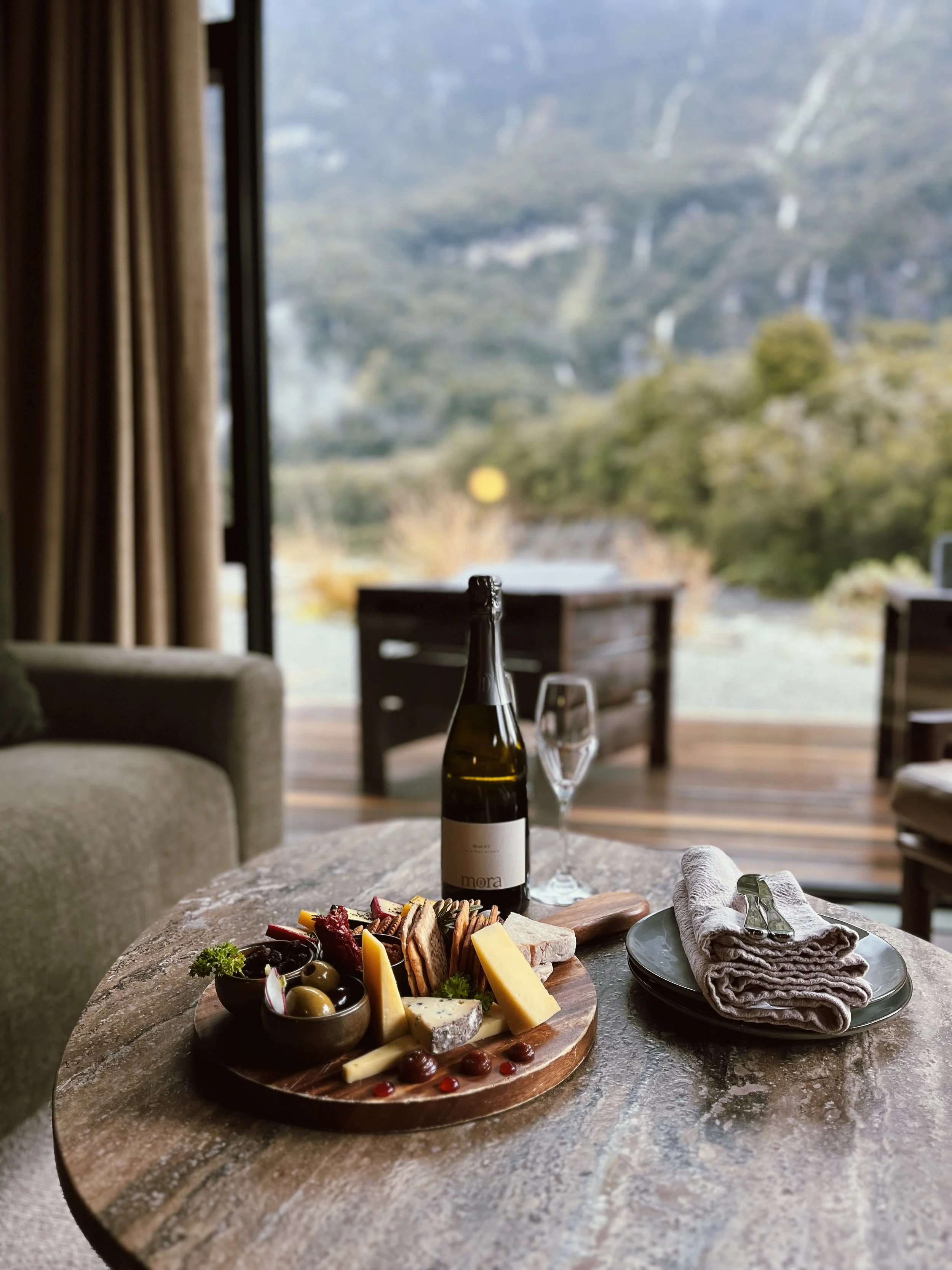 A table with a cheese and charcuterie platter, a bottle of wine, wine glass, napkin, and cutlery, set in a cozy room with a view of mountains and trees through large windows.