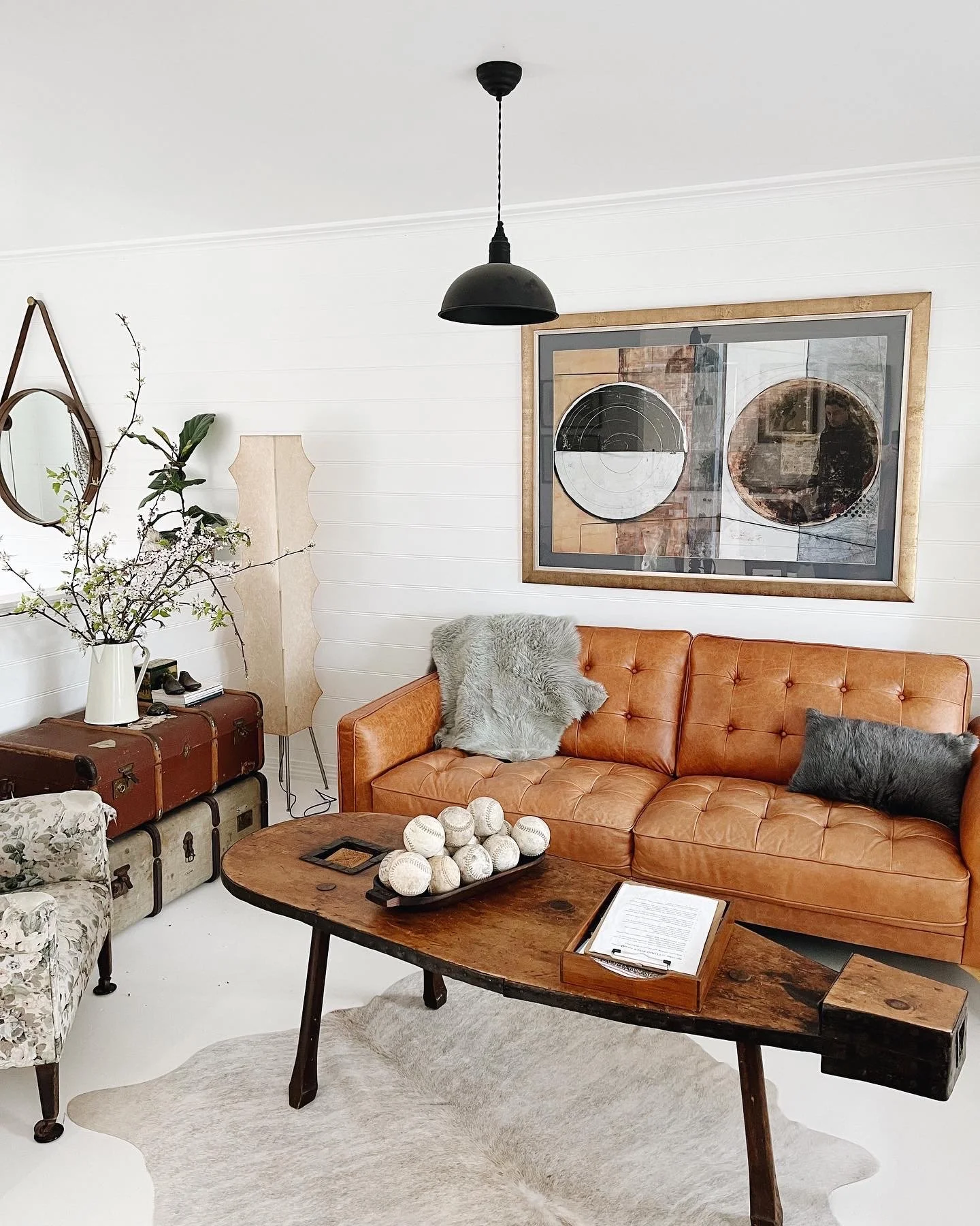 Living room with white paneled walls, a leather sofa with plush pillows, a wooden coffee table with decorative balls, vintage trunks, a tall beige floor lamp, a branch with flowers in a white pitcher, framed abstract artwork on the wall, and a black 