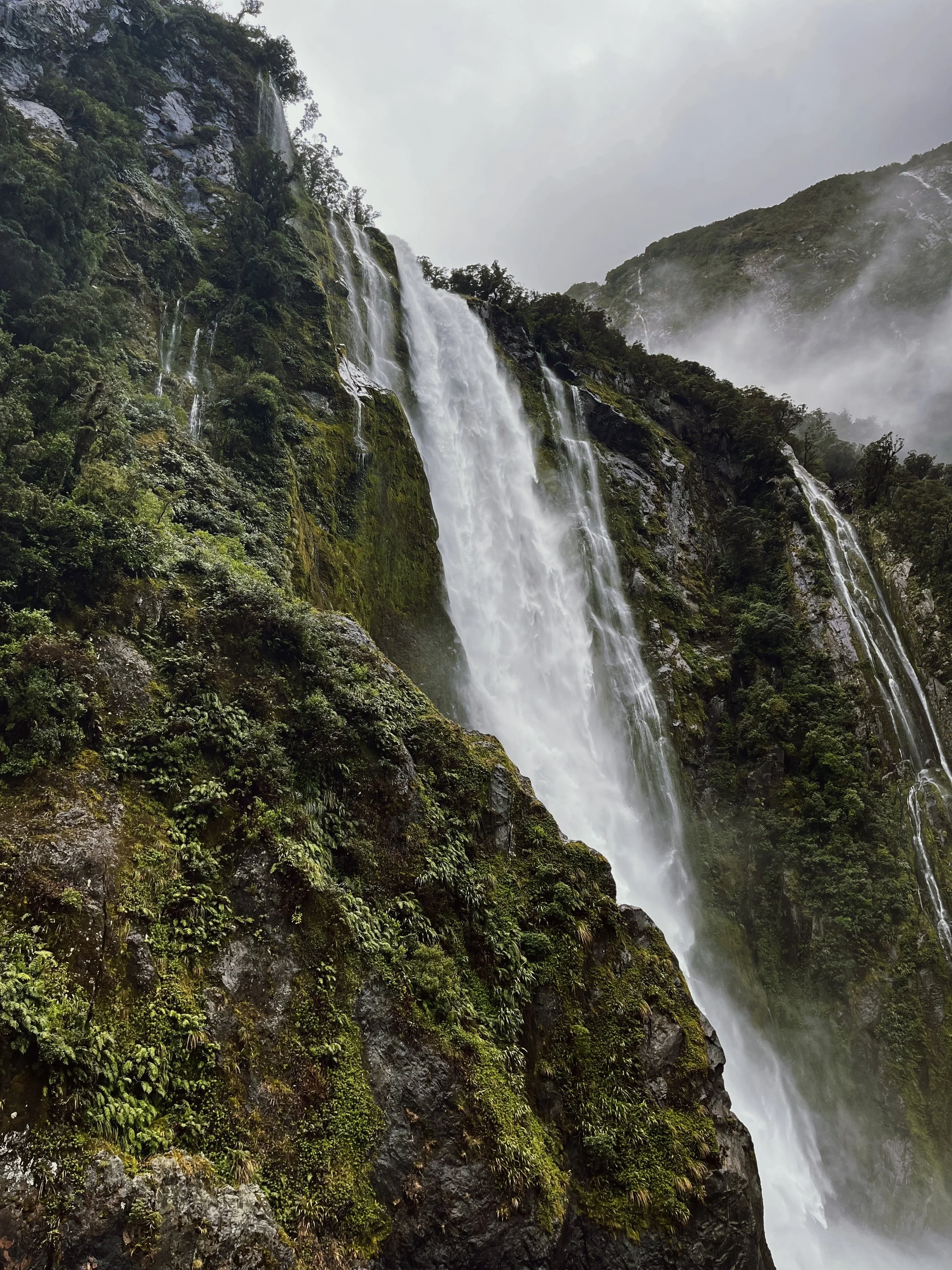 A tall waterfall cascading down a moss-covered rocky mountainside surrounded by lush green trees and vegetation, with mist rising at the base.