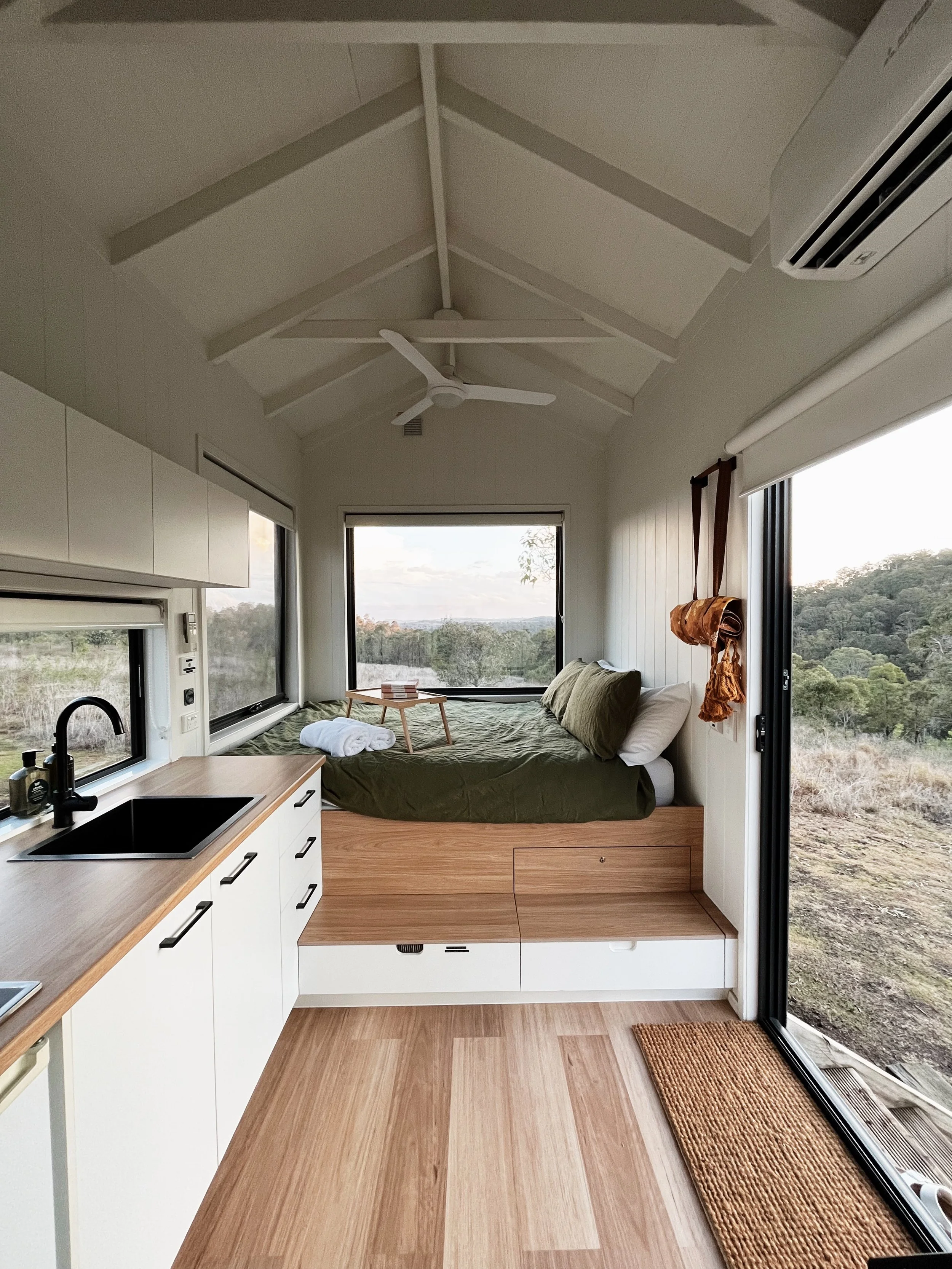 Interior view of a tiny home or camper with a small kitchen area and a bed near a large window, overlooking a landscape with trees and hills.