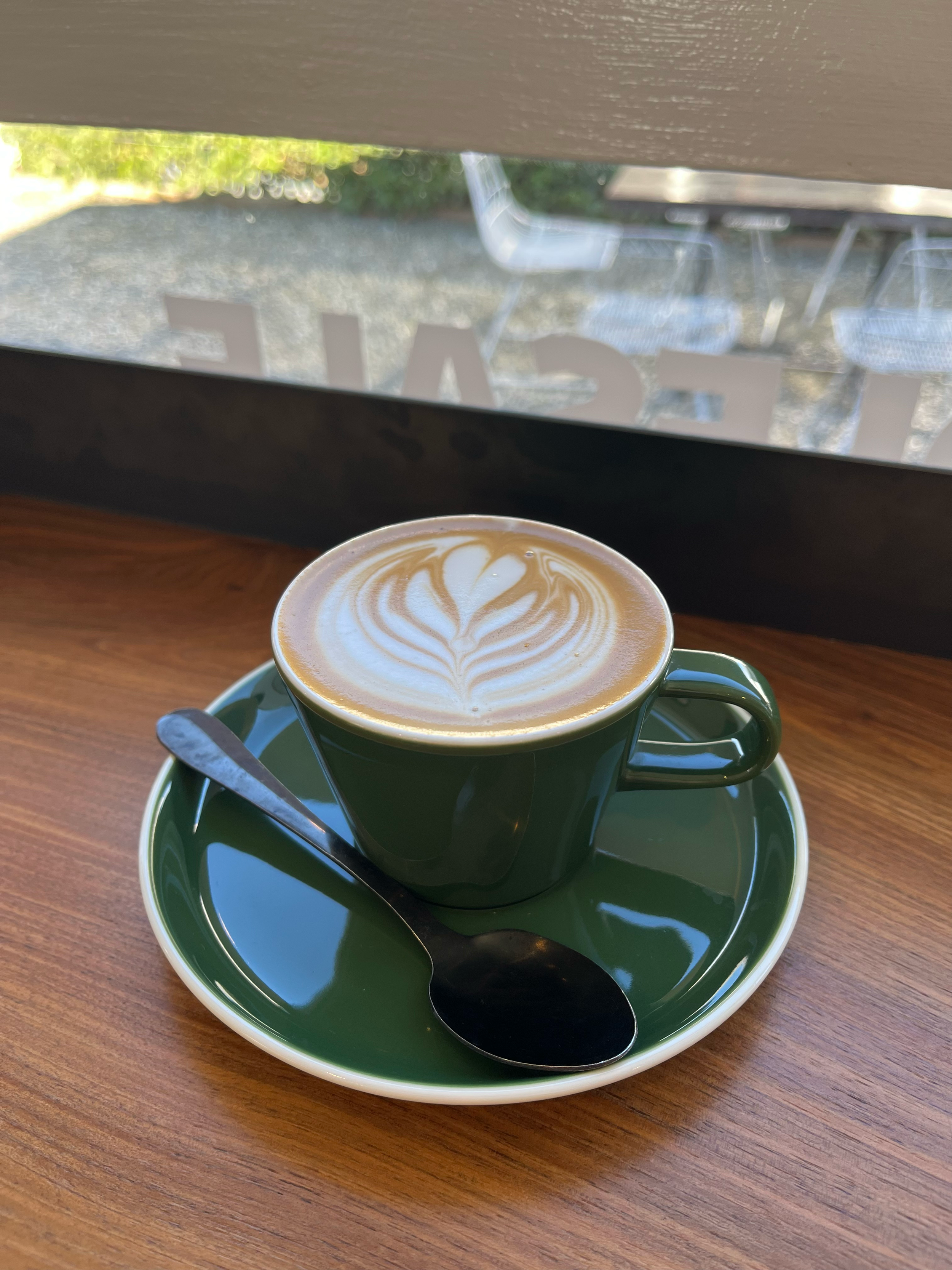 A cup of coffee with latte art on a green saucer, a spoon, on a wooden table near a window with outdoor chairs visible in the background.