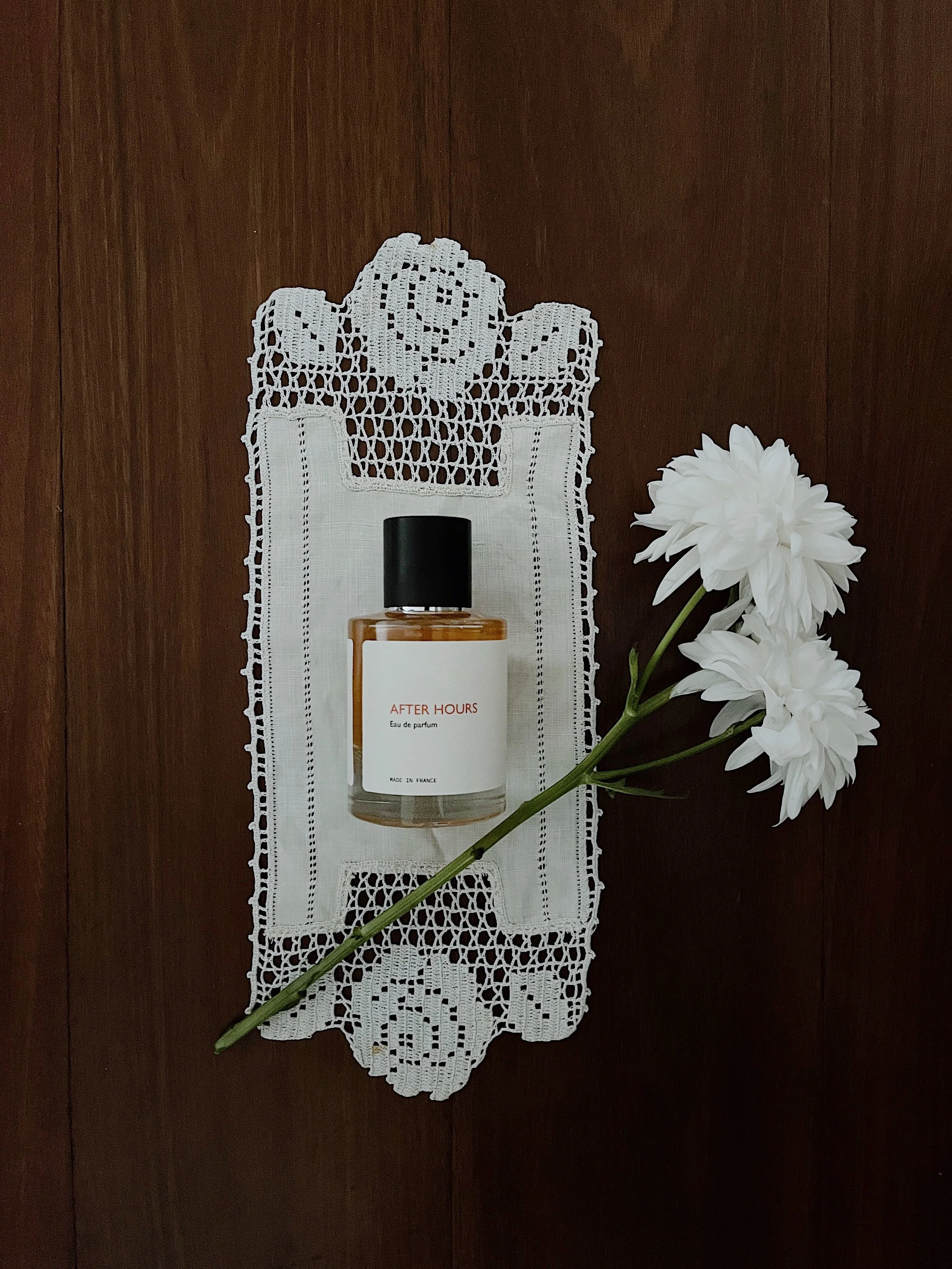 A white doily with lace patterns is placed on a dark wooden surface. On top of the doily, there is a bottle of perfume labeled "AFTER HOURS Eau de parfum MADE IN FRANCE" with a black cap. To the right of the doily, there is a branch of white flowers 