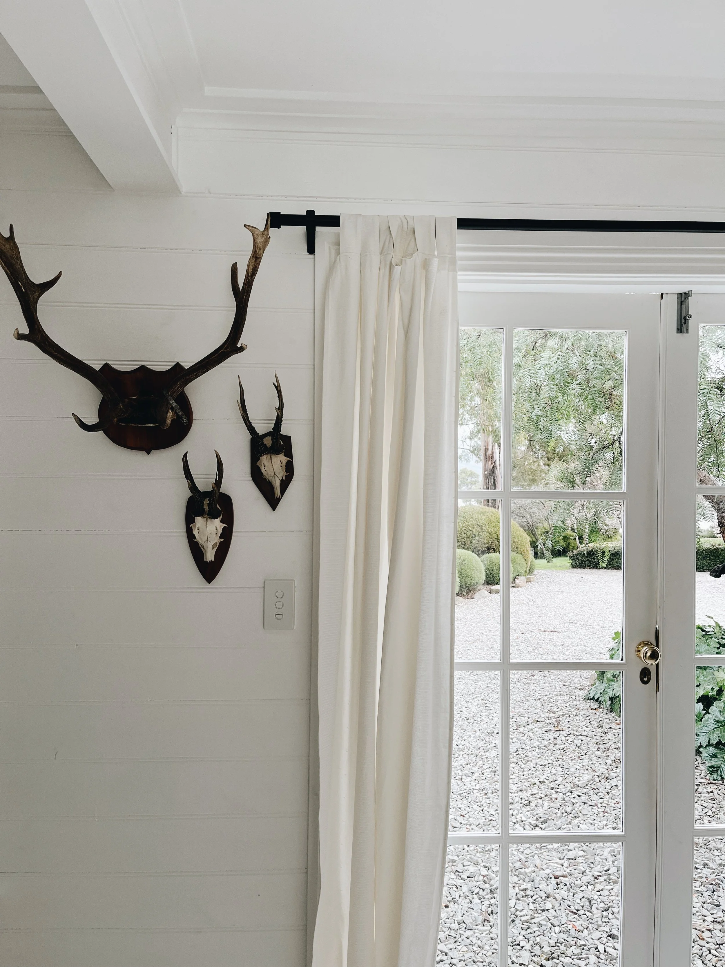 View of a glass door leading to a gravel garden with bushes, trees, and sky outside, with a white curtain on a black rod and mounted animal skulls with antlers on the white wall inside.