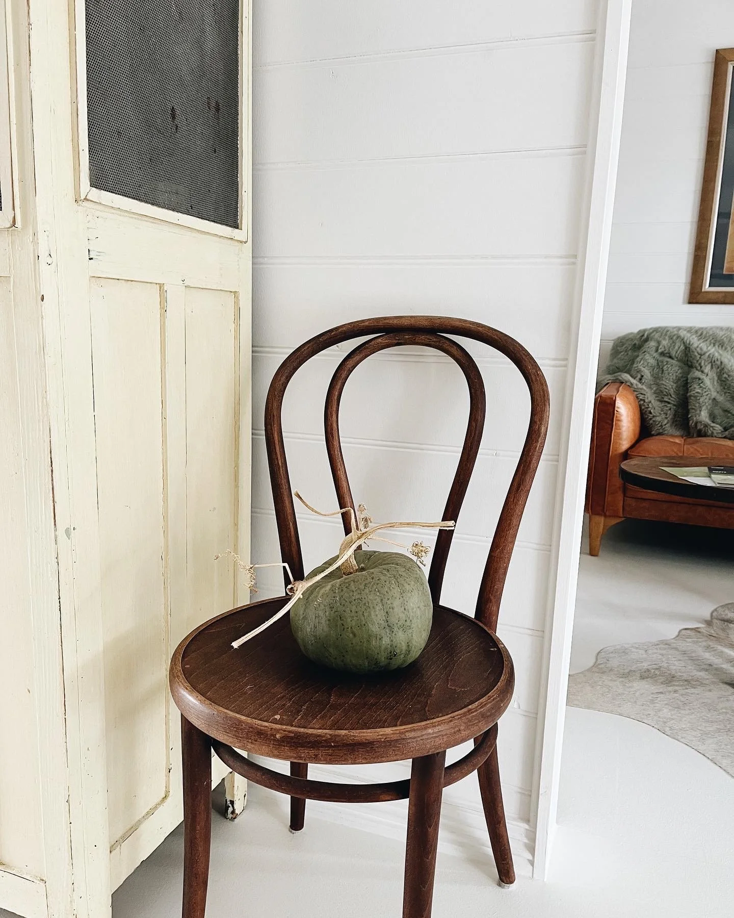 A wooden chair with a round seat holds a green pumpkin with a dried stem inside a room with white walls and a partial view of a sofa and picture frame.