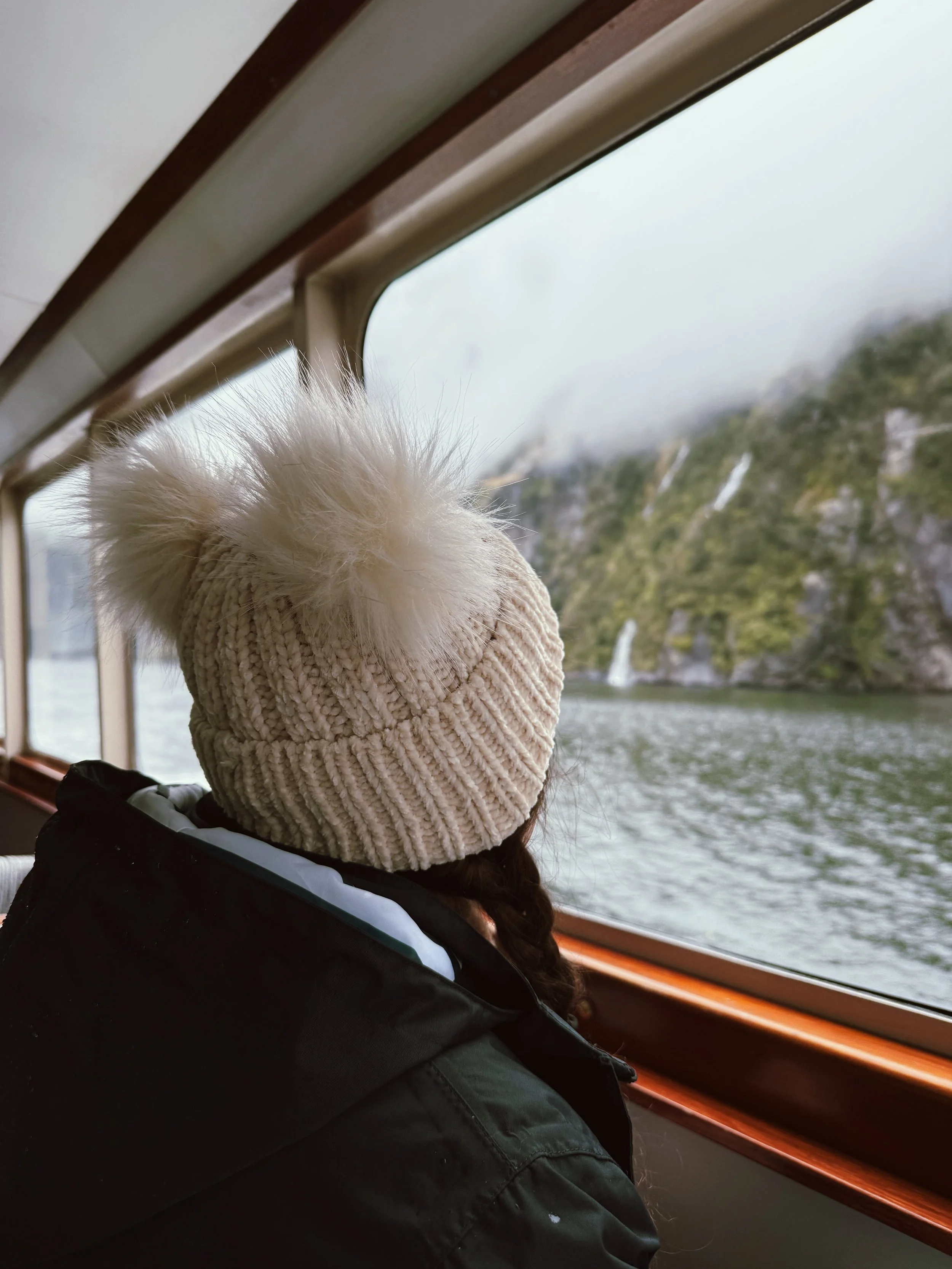 A woman wearing a knitted beige beanie with a fluffy pom-pom on top, sitting by a window on a boat, looking outside at a body of water and a rocky hillside with waterfalls.