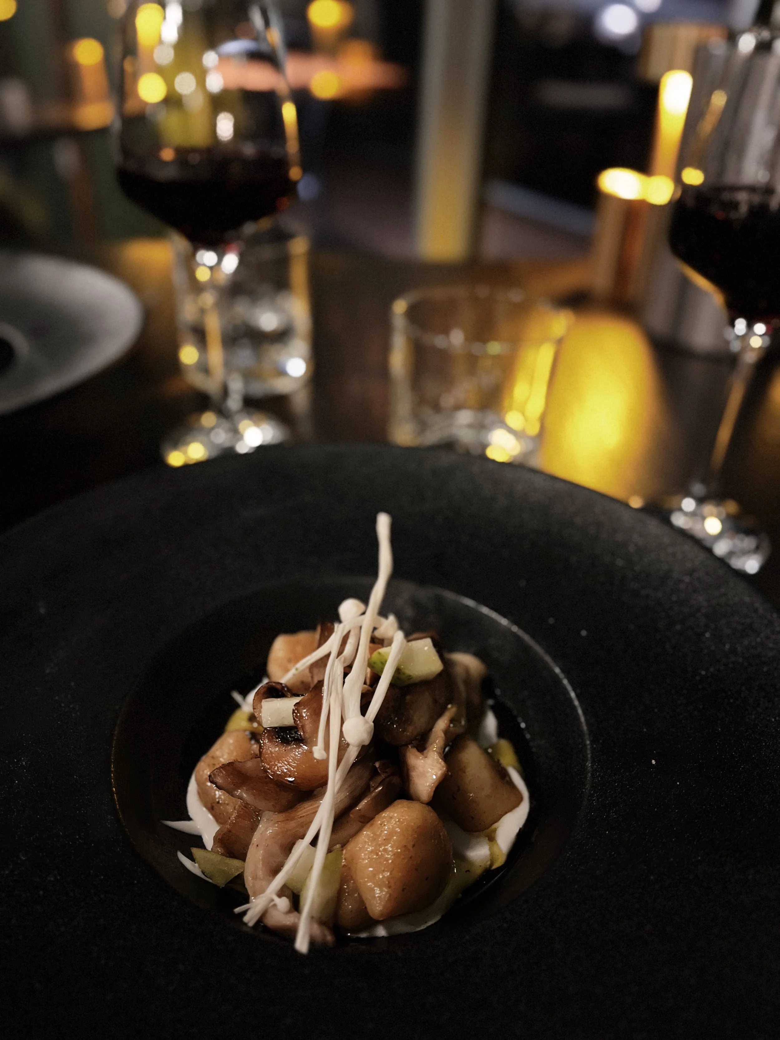 A small dish of cooked mushrooms, garnished with bean sprouts and chopped green onions, served in a black bowl on a dark table with a blurry background of wine glasses and dim lighting.