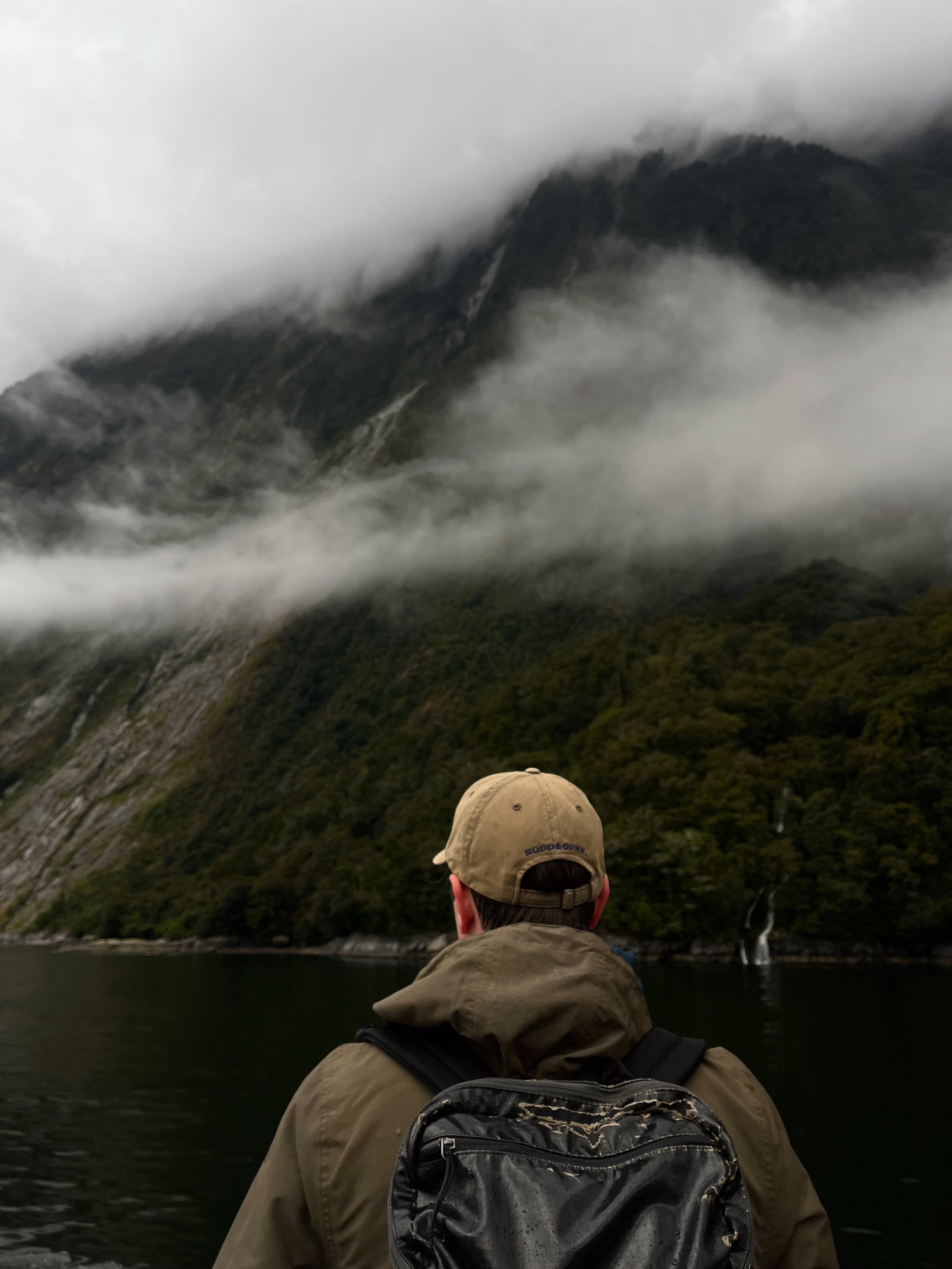 A person with a backpack and beige hat observing a misty mountain with waterfalls.