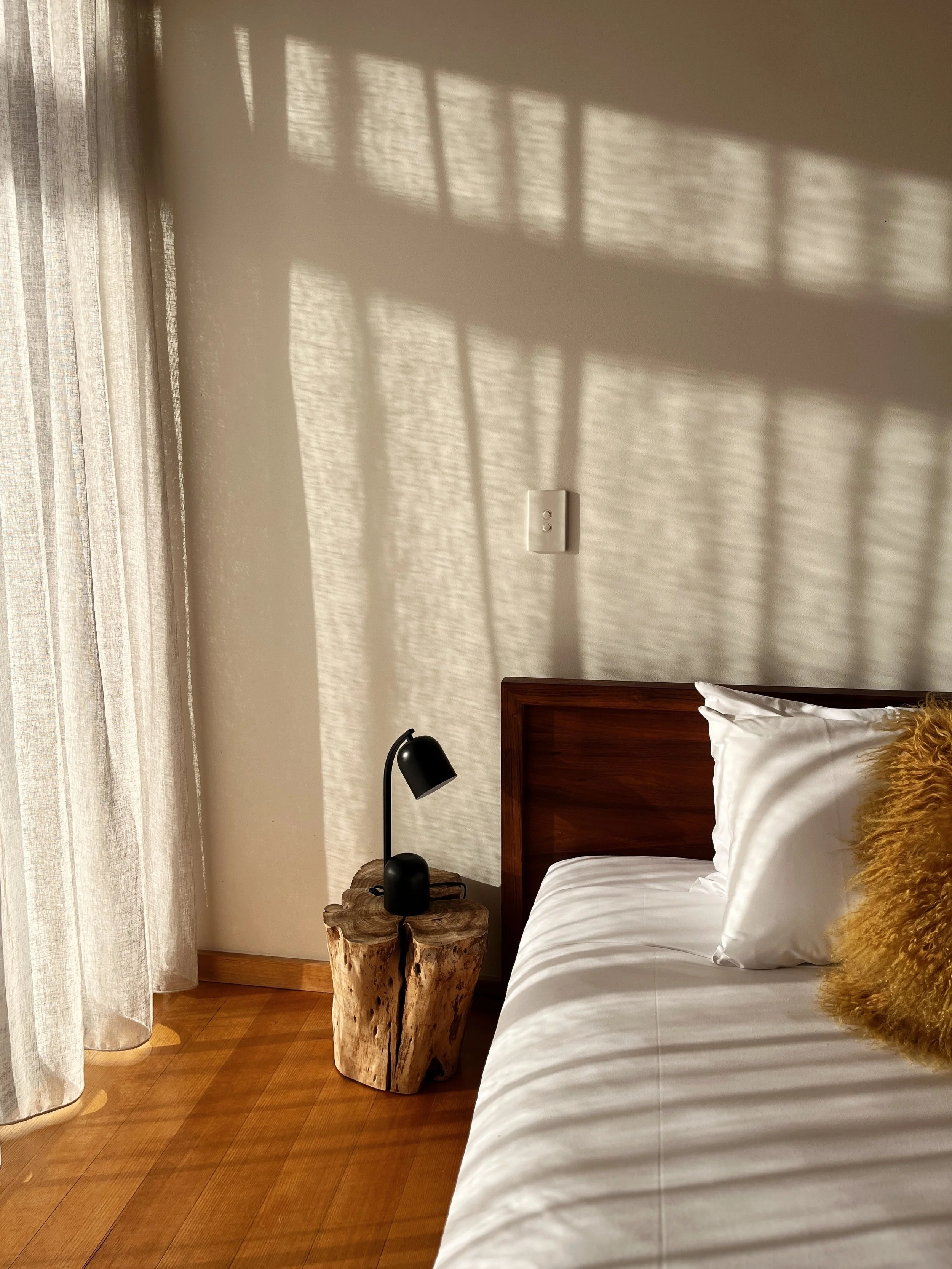 A bedroom with a bed featuring white linens and a gold textured pillow. A wooden headboard, a side table made from a natural wood slab, a black desk lamp, and a black alarm clock are visible. Light filters through sheer curtains, casting a shadow on 