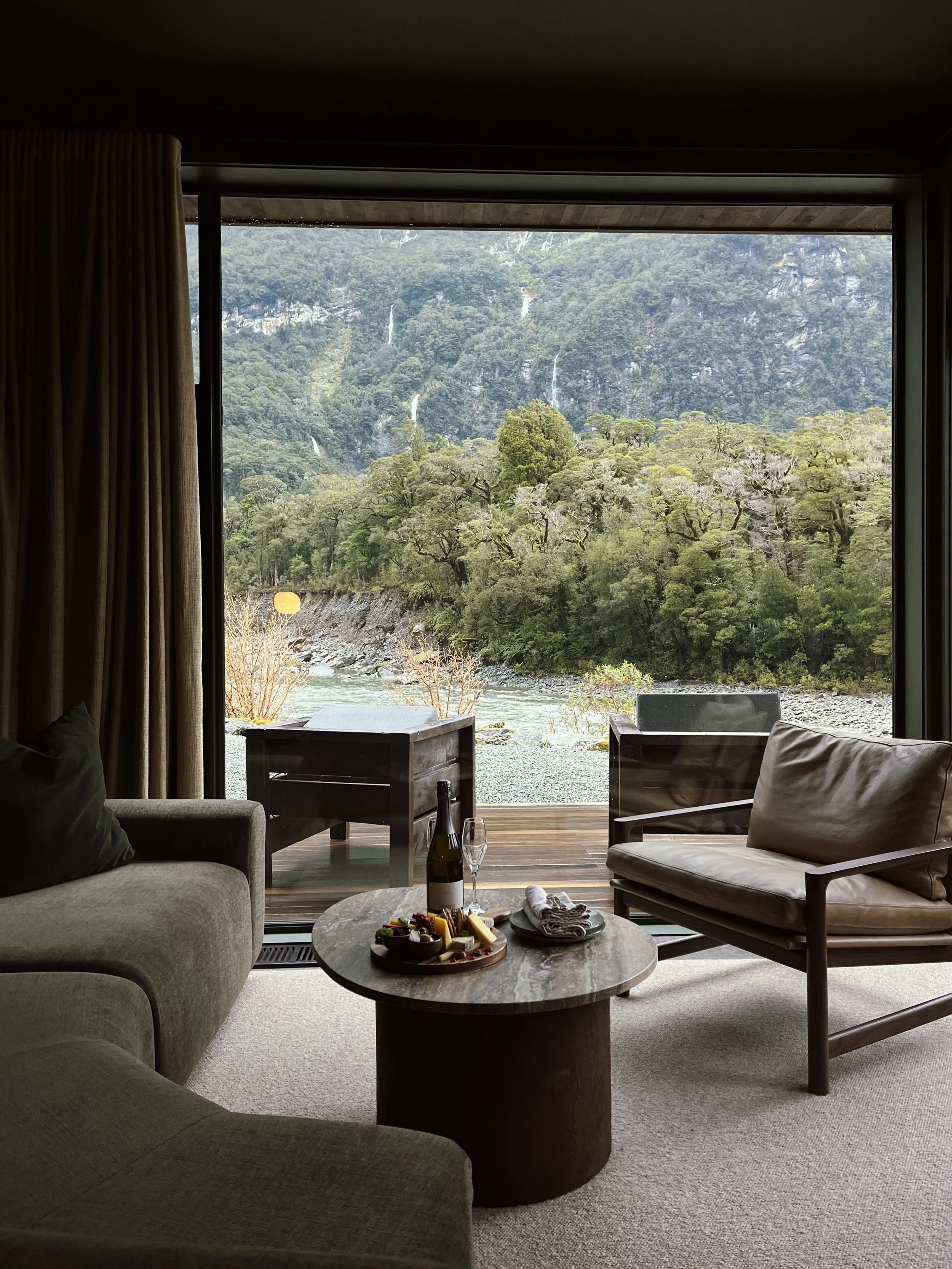 Living room with a large window overlooking a scenic mountain landscape with trees and waterfalls, furniture includes a couch, armchair, and a coffee table with wine, cheese, and snacks.