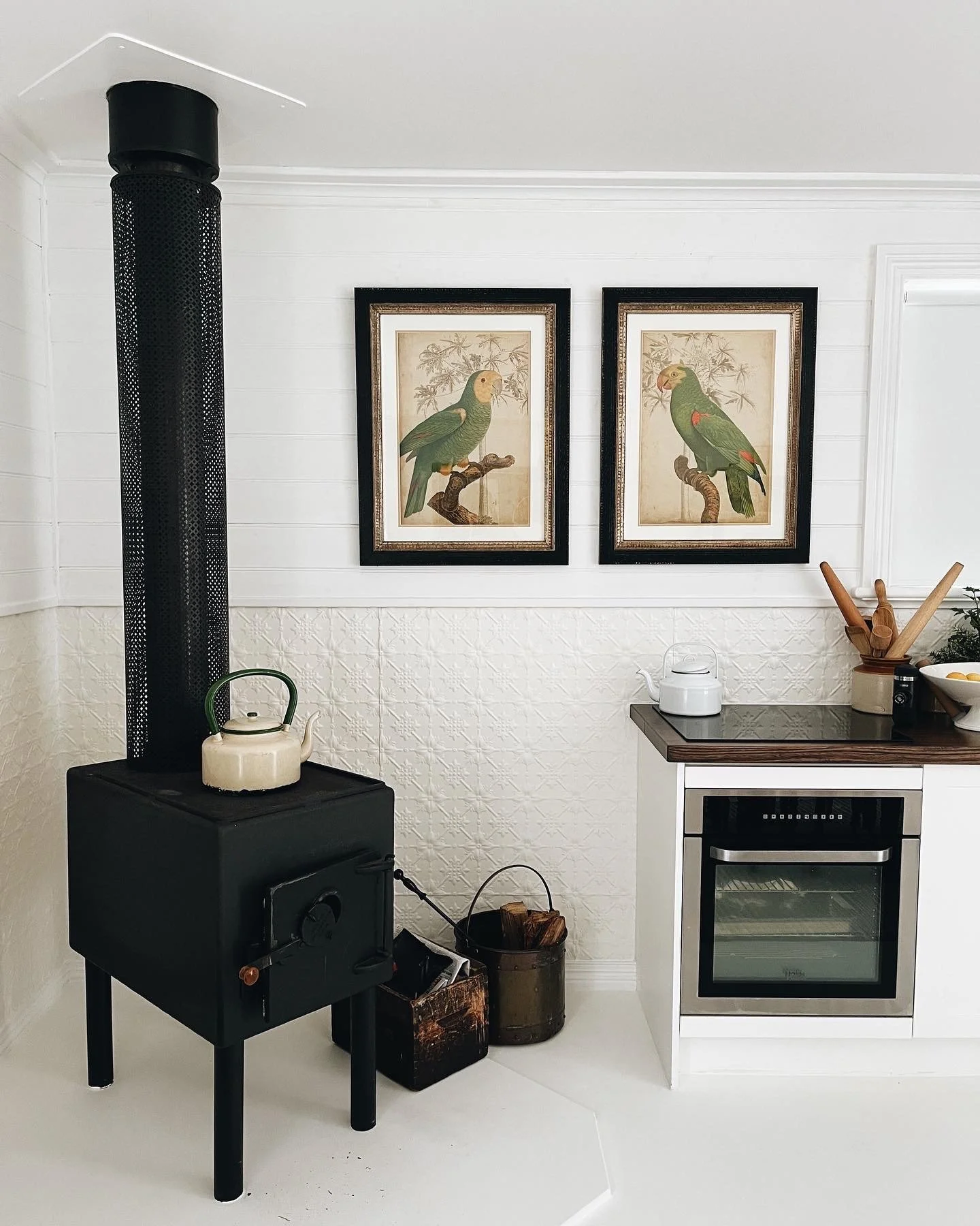 A cozy kitchen scene featuring a wood stove with a kettle on top, framed bird artwork on the wall, and a small built-in oven with wooden utensils in a holder. There are also decorative vintage containers and a small potted plant on the counter.