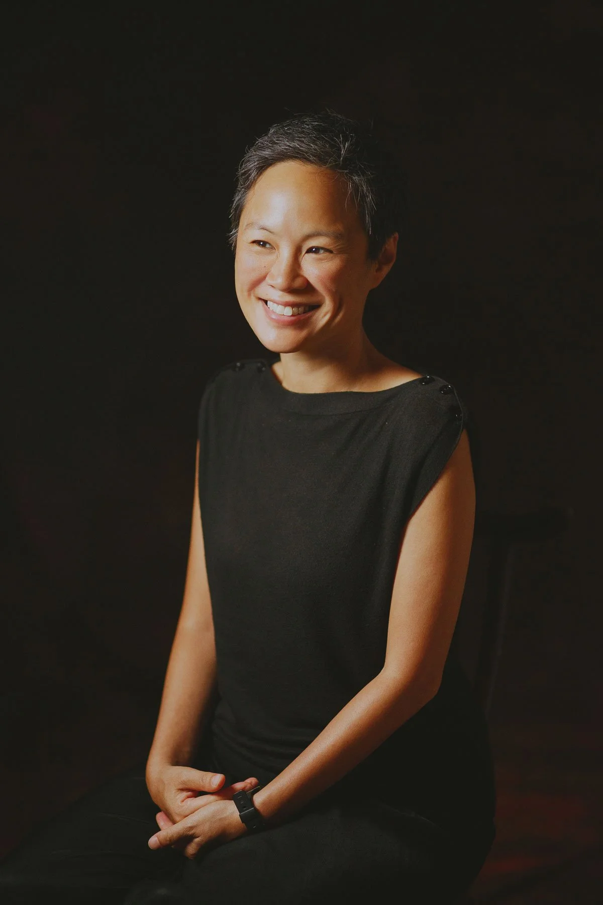 Portrait of a woman with short dark hair wearing a sleeveless black top, sitting with hands clasped, smiling against a dark background.