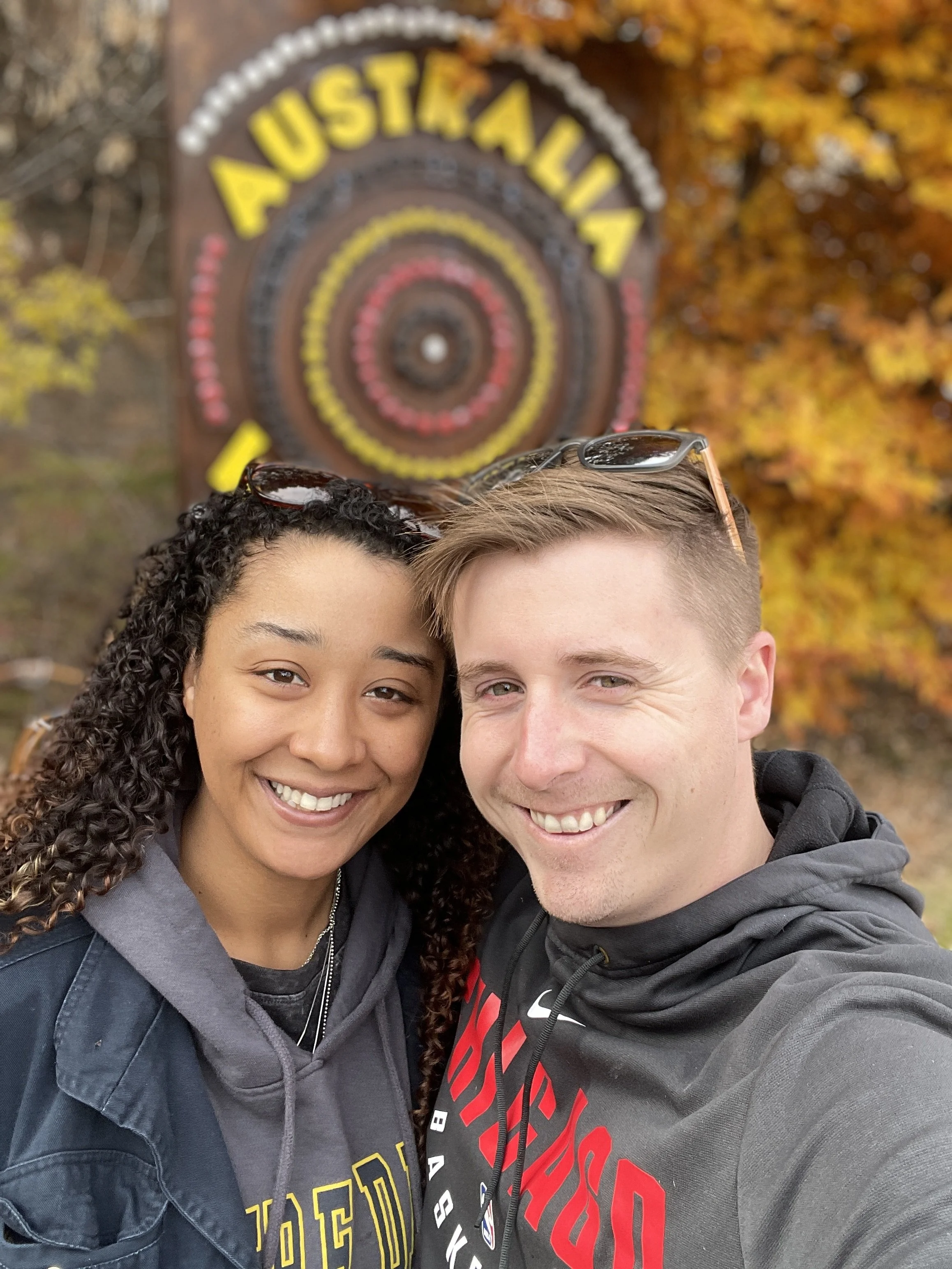 Two smiling people taking a selfie outdoors with fall foliage in the background, a woman with curly dark hair and glasses on her head, and a man with short light hair wearing a gray jacket.