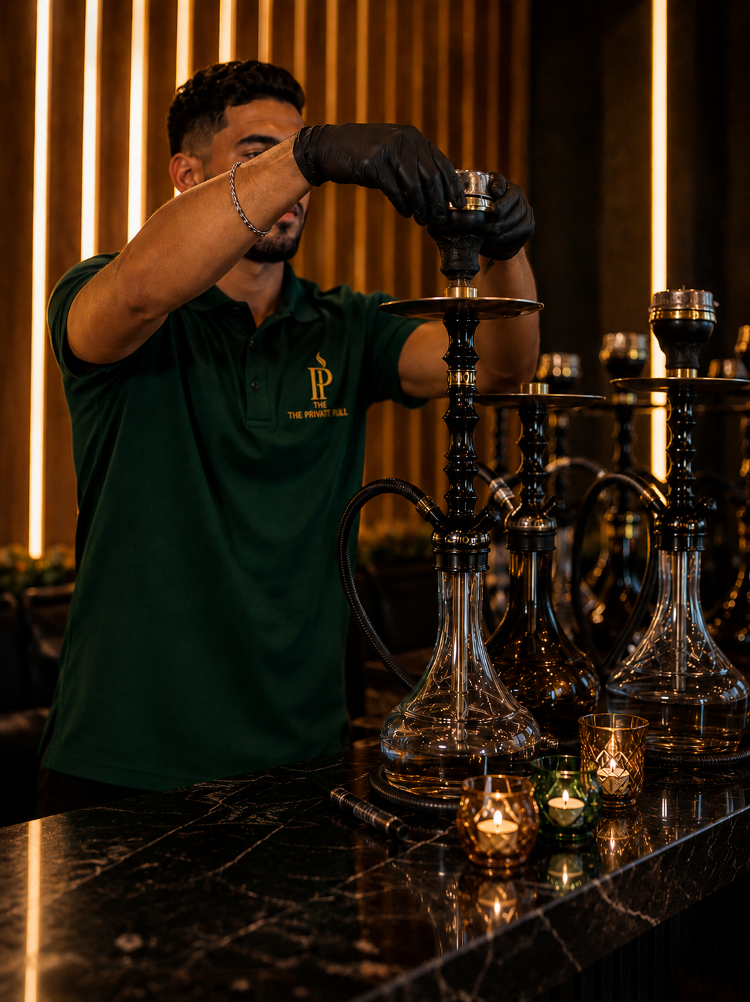 A man in a green polo shirt and black gloves preparing a hookah on a black marble table, with candle holders and a wooden wall in the background.