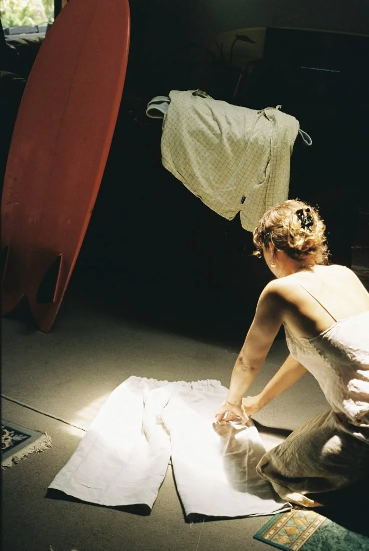 A woman with curly hair is kneeling on the floor, folding a white pair of pants. There is a surfboard, some folded clothes, and a laptop nearby in a dimly lit room.