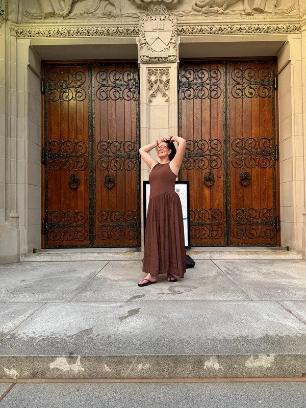 A woman in a brown dress and sandals standing in front of large, ornate wooden gates with black wrought iron scrollwork, on a stone building with decorative stone carvings, smiling and stretching with hands behind her head.