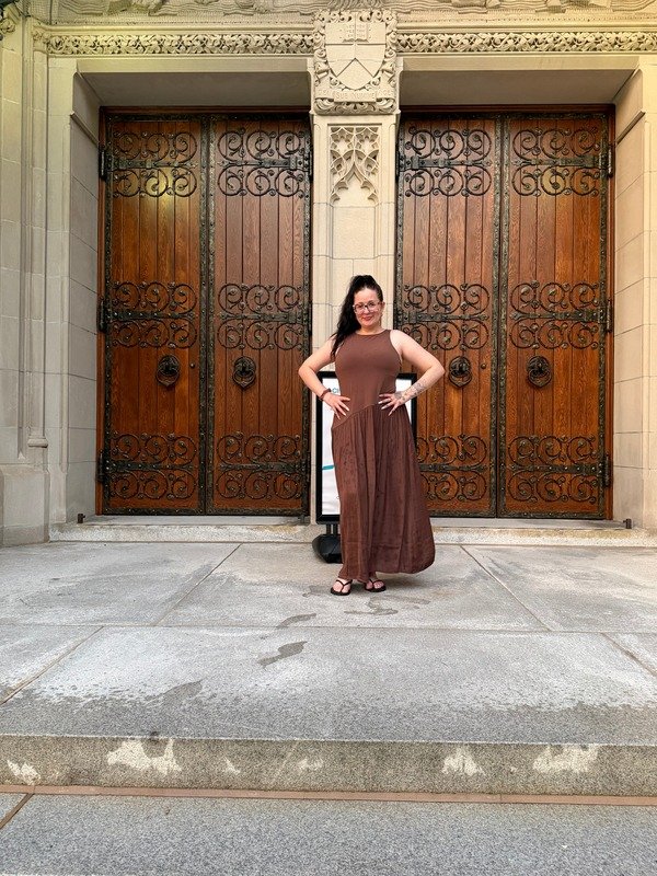 A woman in a brown dress standing in front of large wooden doors with decorative wrought iron details, on a stone building with ornate architectural features.