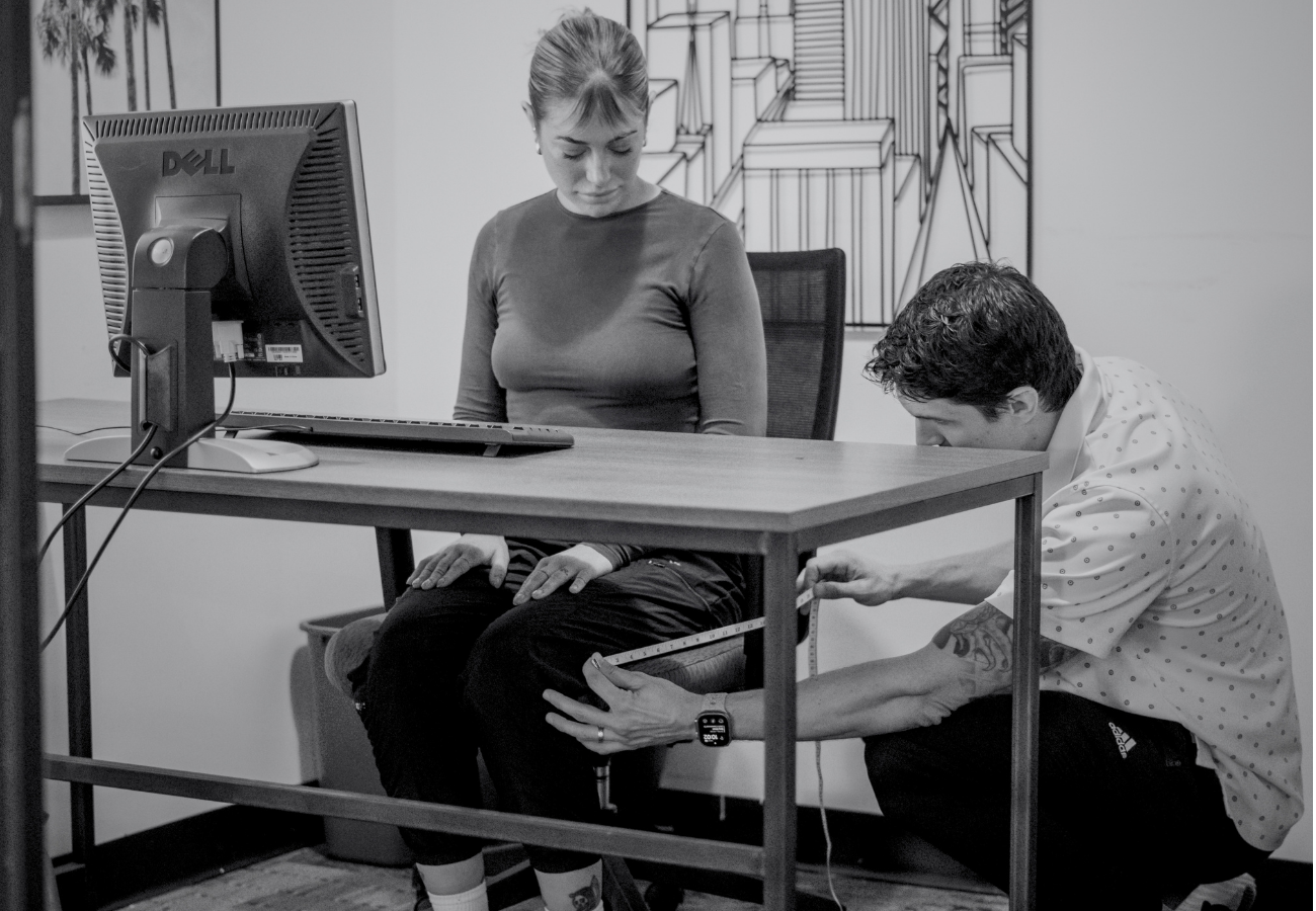 A woman sitting at a desk with a computer looks down as a man measures her thigh and ankle using a measuring tape in an office setting.