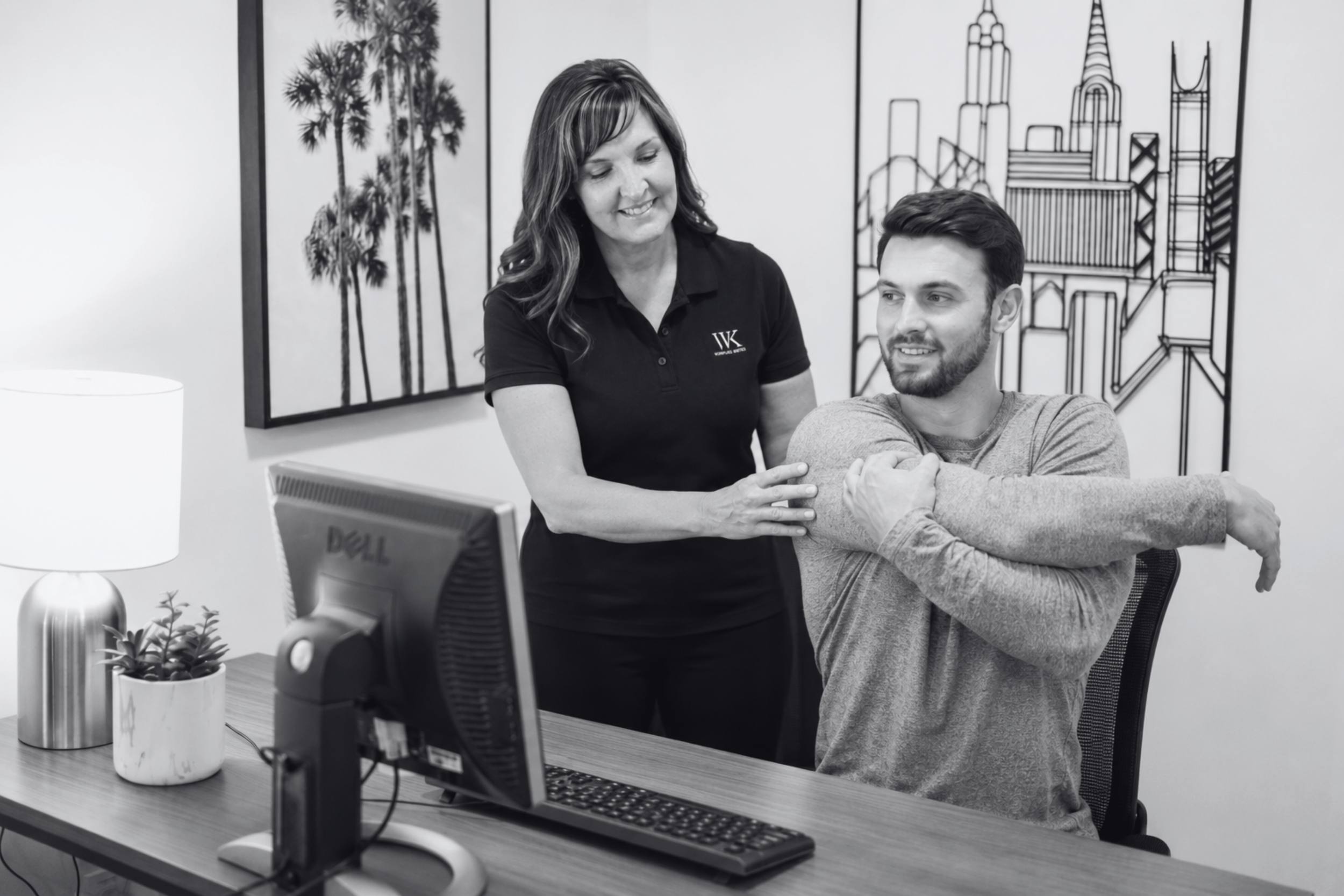 A woman is sitting at a desk showing her arm to a smiling man, who is seated. The woman has long hair and is wearing a black polo shirt with a logo. The man has short hair and a beard, and is wearing a gray sweater. The desk has a computer monitor, keyboard, mouse, a small potted plant, and a lamp. In the background, there are framed artwork of trees and a cityscape.