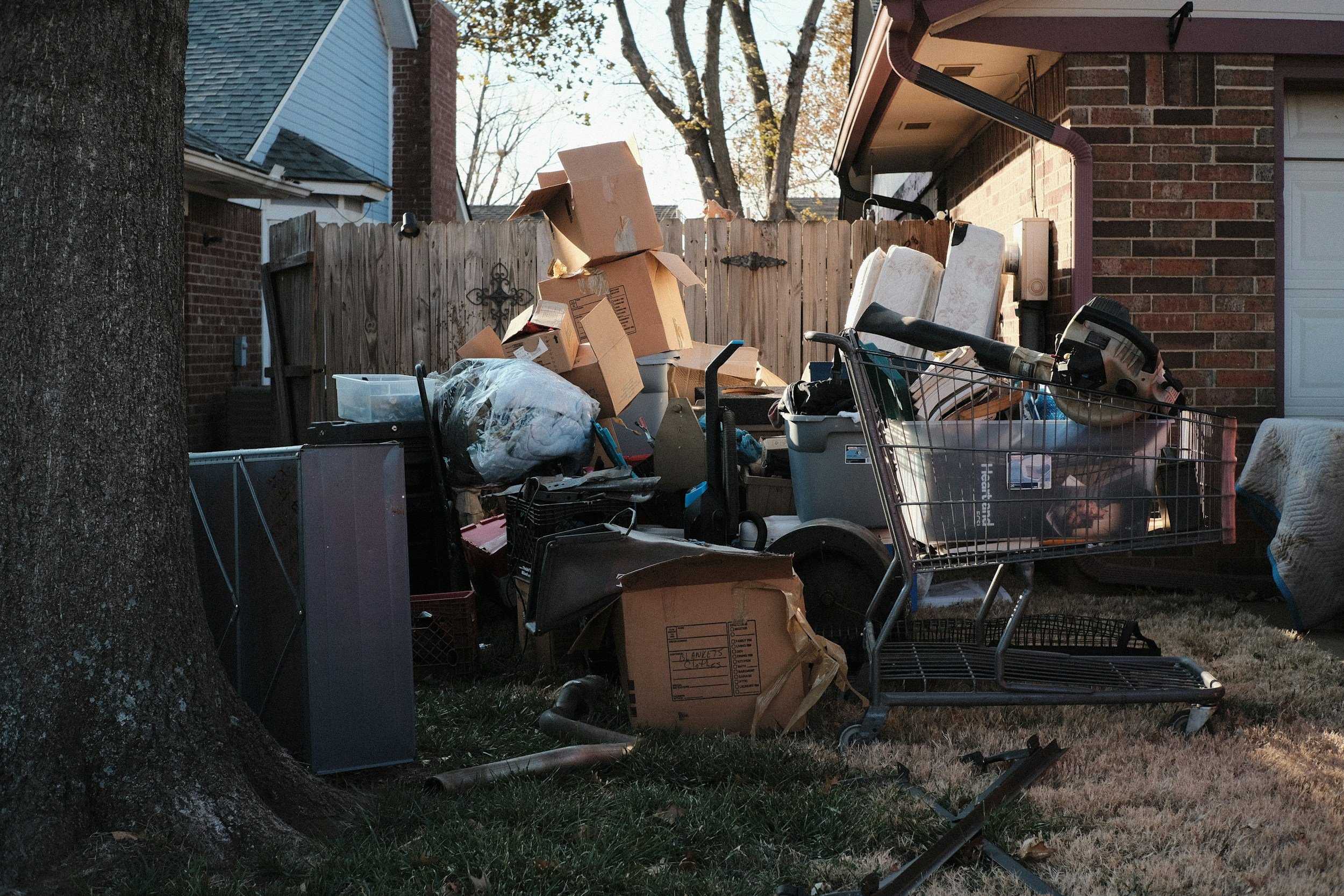 Pile of discarded household items and cardboard boxes outside in a yard, next to a brick house and fence, with trees in the background.