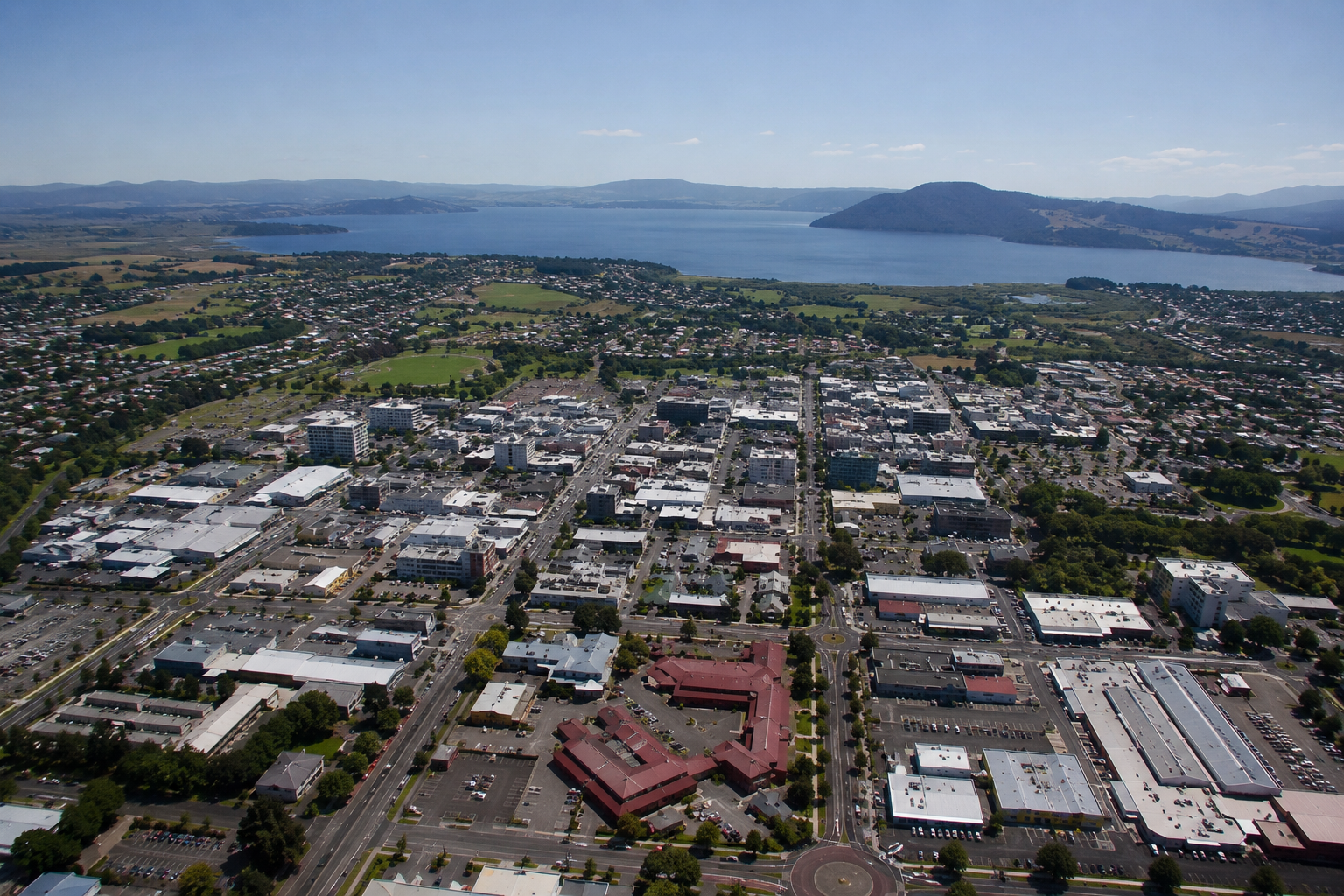 Aerial view of a city with a lake in the background, featuring a mix of commercial and residential buildings, green spaces, and parking lots under a clear blue sky.