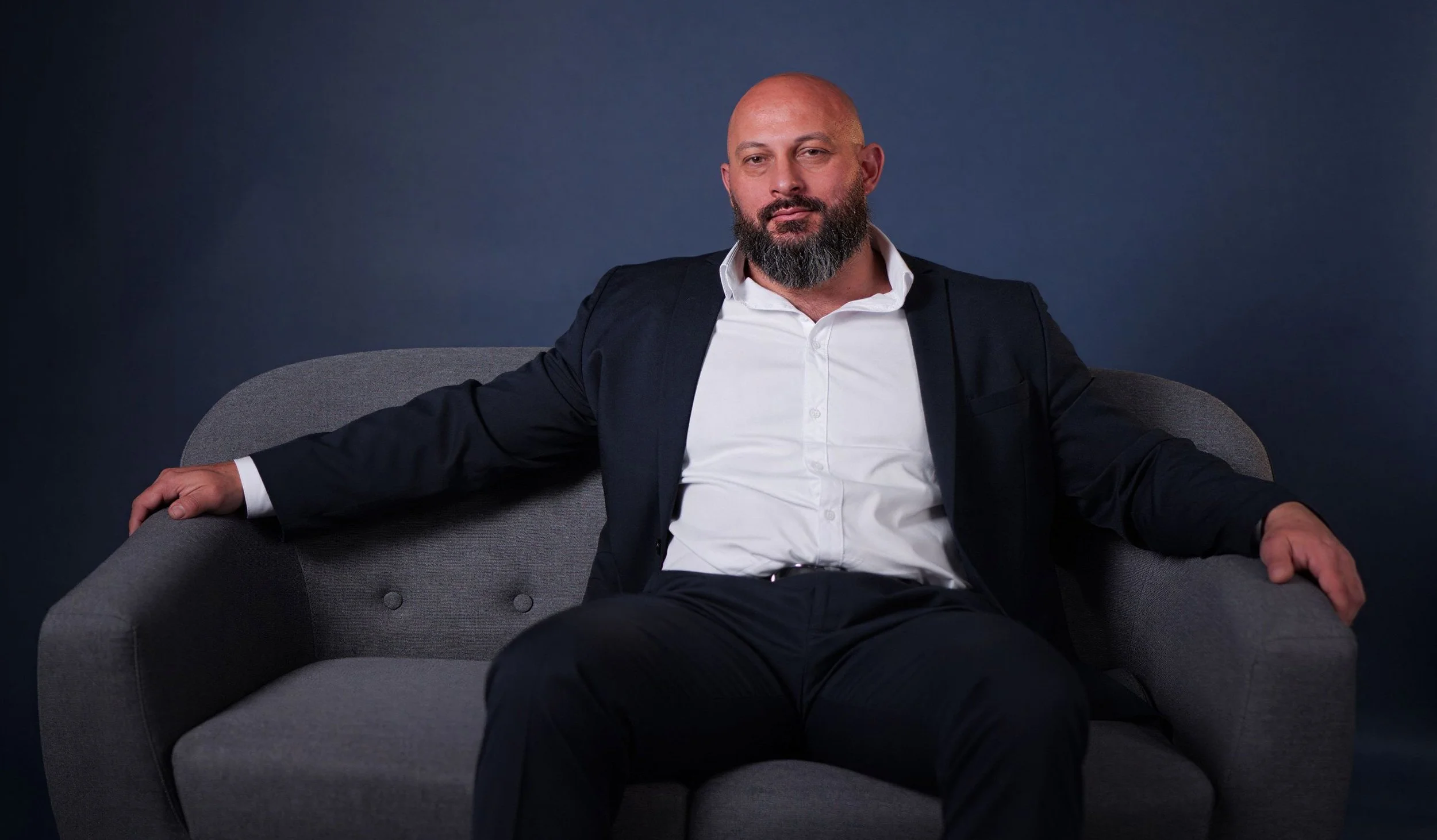 A confident man with a beard, bald head, wearing a white dress shirt and dark blazer, sitting on a gray couch against a dark blue background.