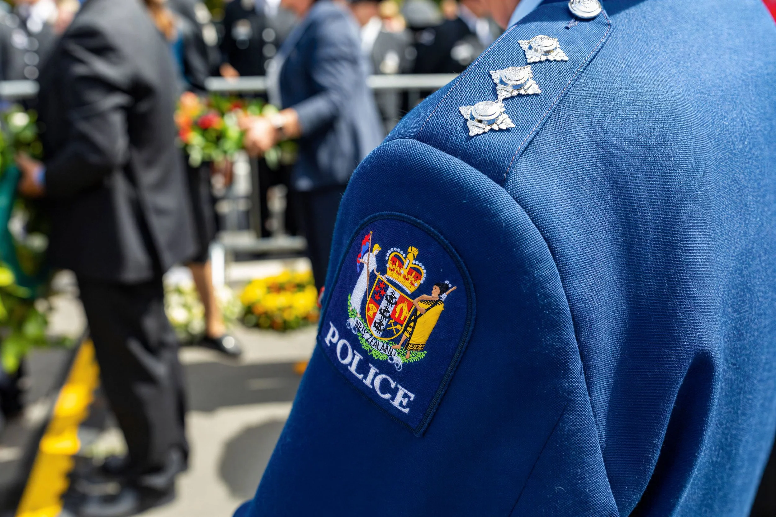 Close-up of a police officer's blue uniform with a shoulder patch that says 'Police' and the New Zealand emblem. Background shows indistinct people, some holding flowers, possibly at a public event or memorial.