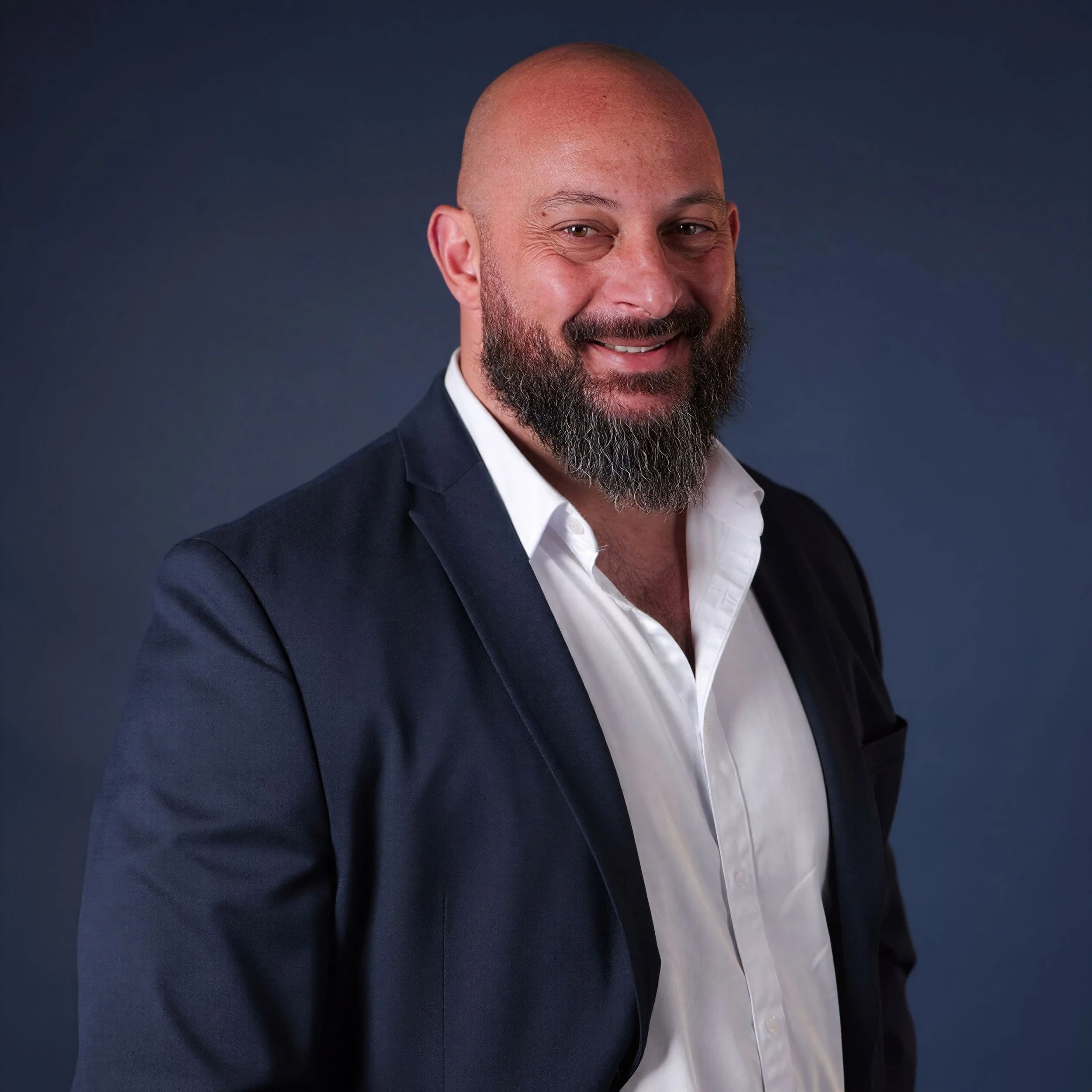 A man with a bald head, beard, and mustache wearing a dark suit jacket and white shirt, smiling against a dark blue background.