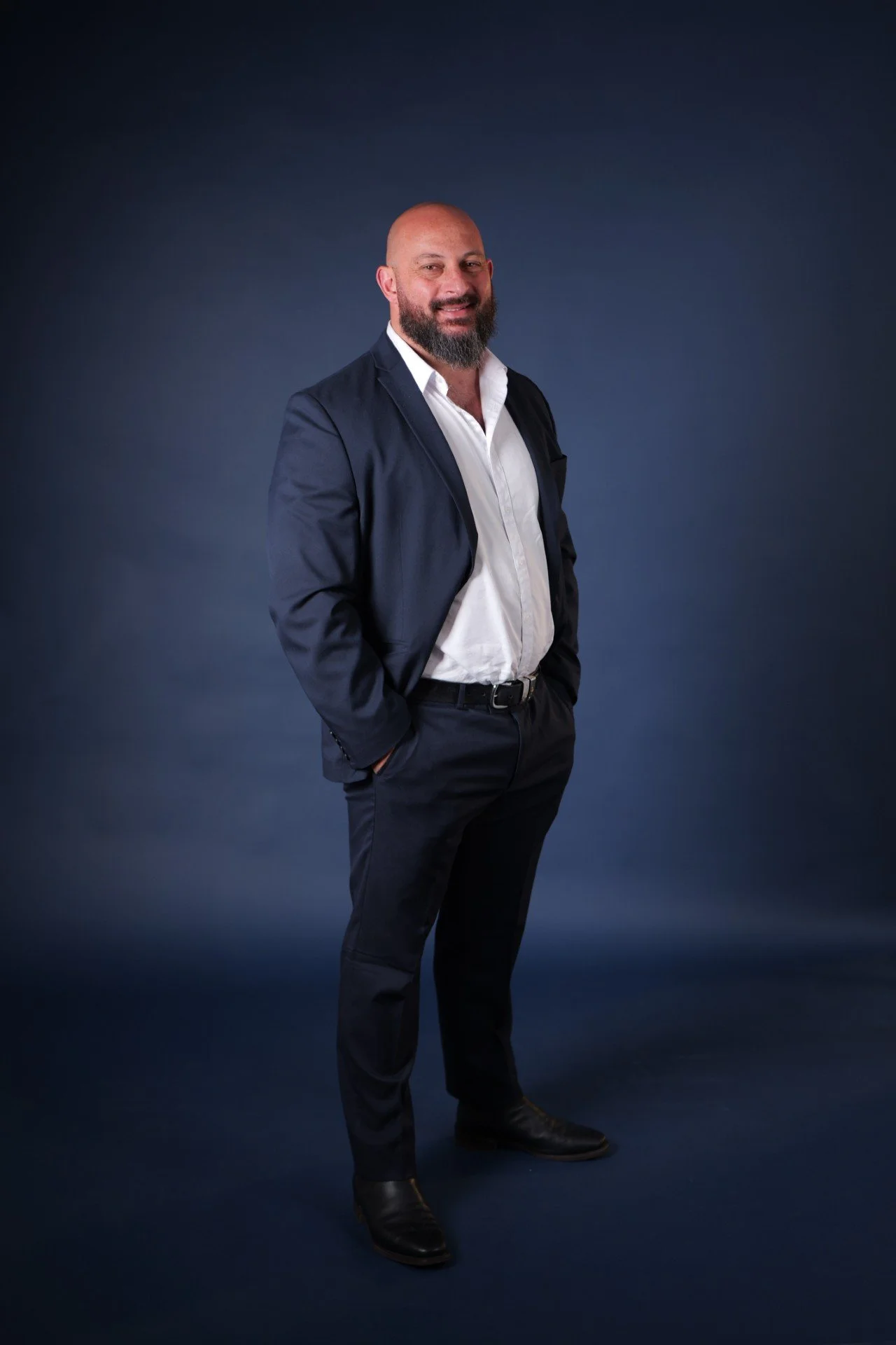 Portrait of a man with a beard wearing a navy suit and white shirt, standing with hands in pockets against a dark blue background.
