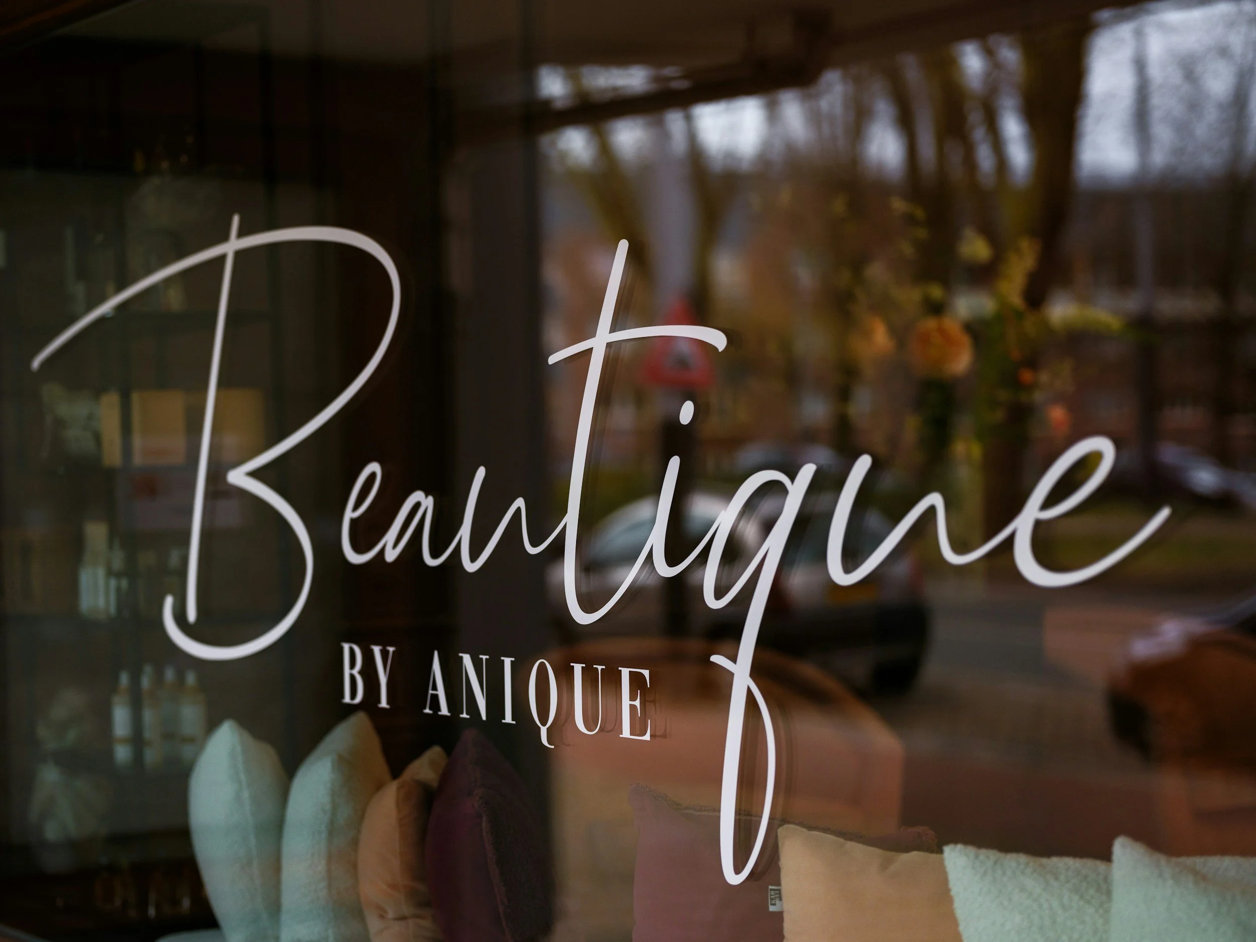A close-up of a glass storefront window with white cursive text that reads 'Breathtaking by Anoufe.' The slide shows an interior with soft pastel-colored treats or items, and a blurred view of trees and a street outside.