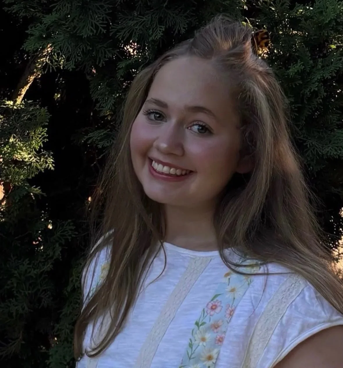 Young woman with long brown hair smiling outdoors in front of green foliage, wearing a white top with floral embroidery.