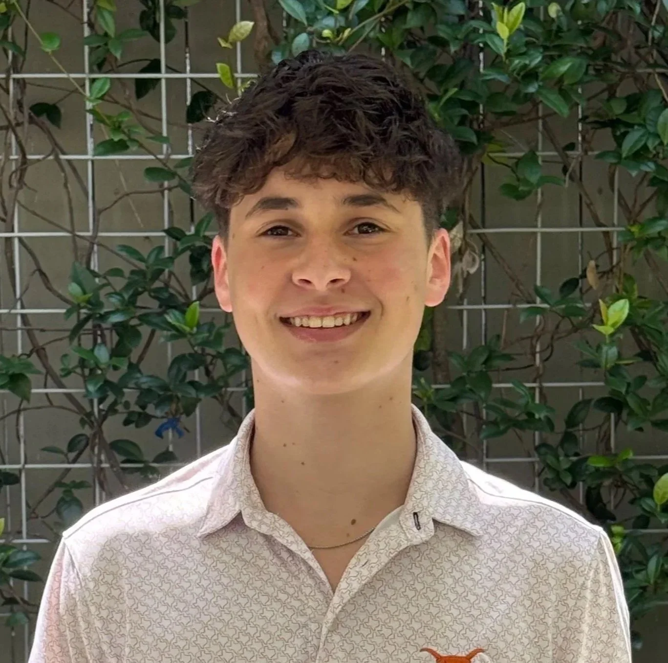 A young man with curly brown hair smiling, standing in front of a metal grid and green leafy plants.