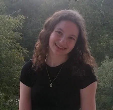 Young woman with curly hair smiling outdoors with greenery in the background.