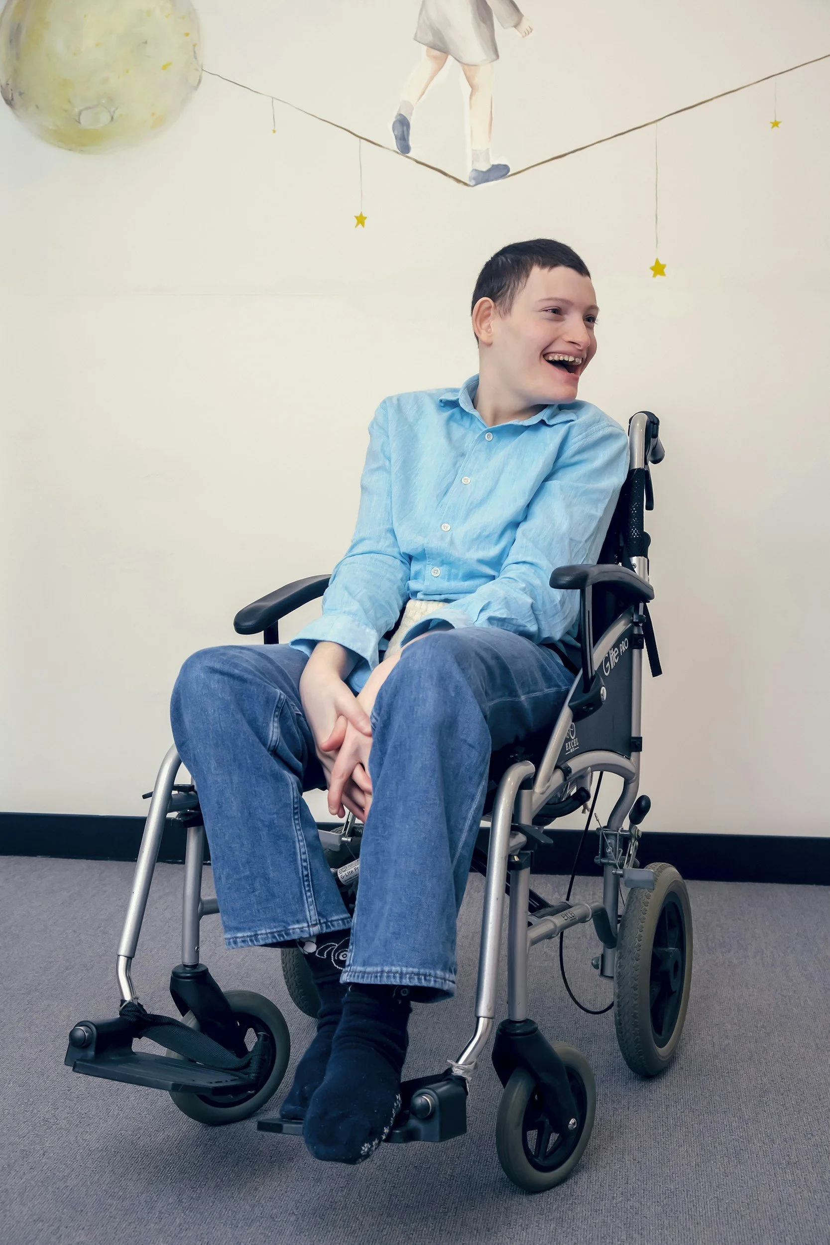 A smiling young man with short dark hair sitting in a wheelchair against a plain wall with a mural of a child walking on a tightrope, stars, and a planet painted on it.