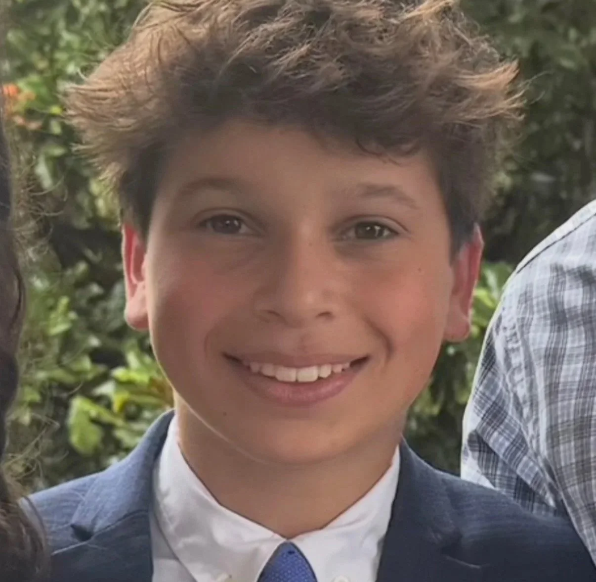 A young man with curly brown hair and light skin, smiling, wearing a blue blazer, white shirt, and blue tie, standing outdoors with greenery in the background.