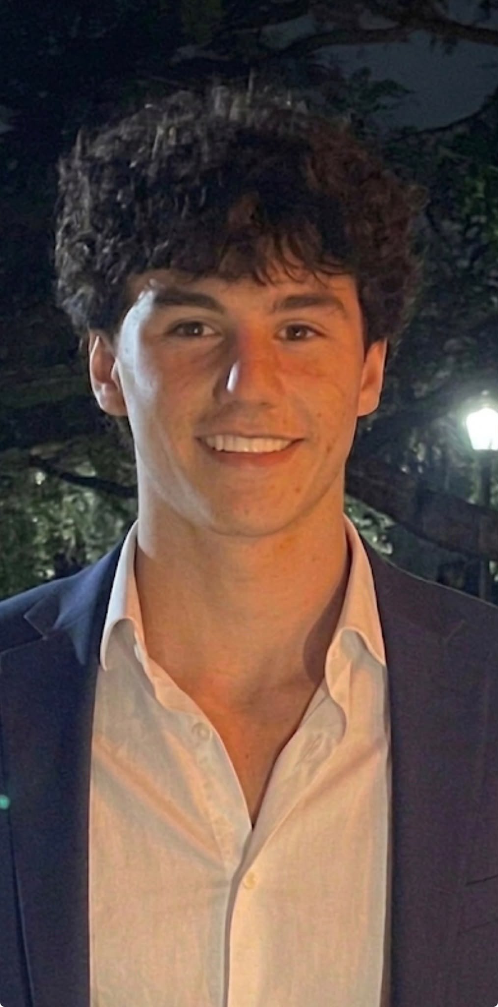 A young man with wavy dark hair, wearing a dark blazer and white shirt, smiling outdoors at night with trees and lights in the background.