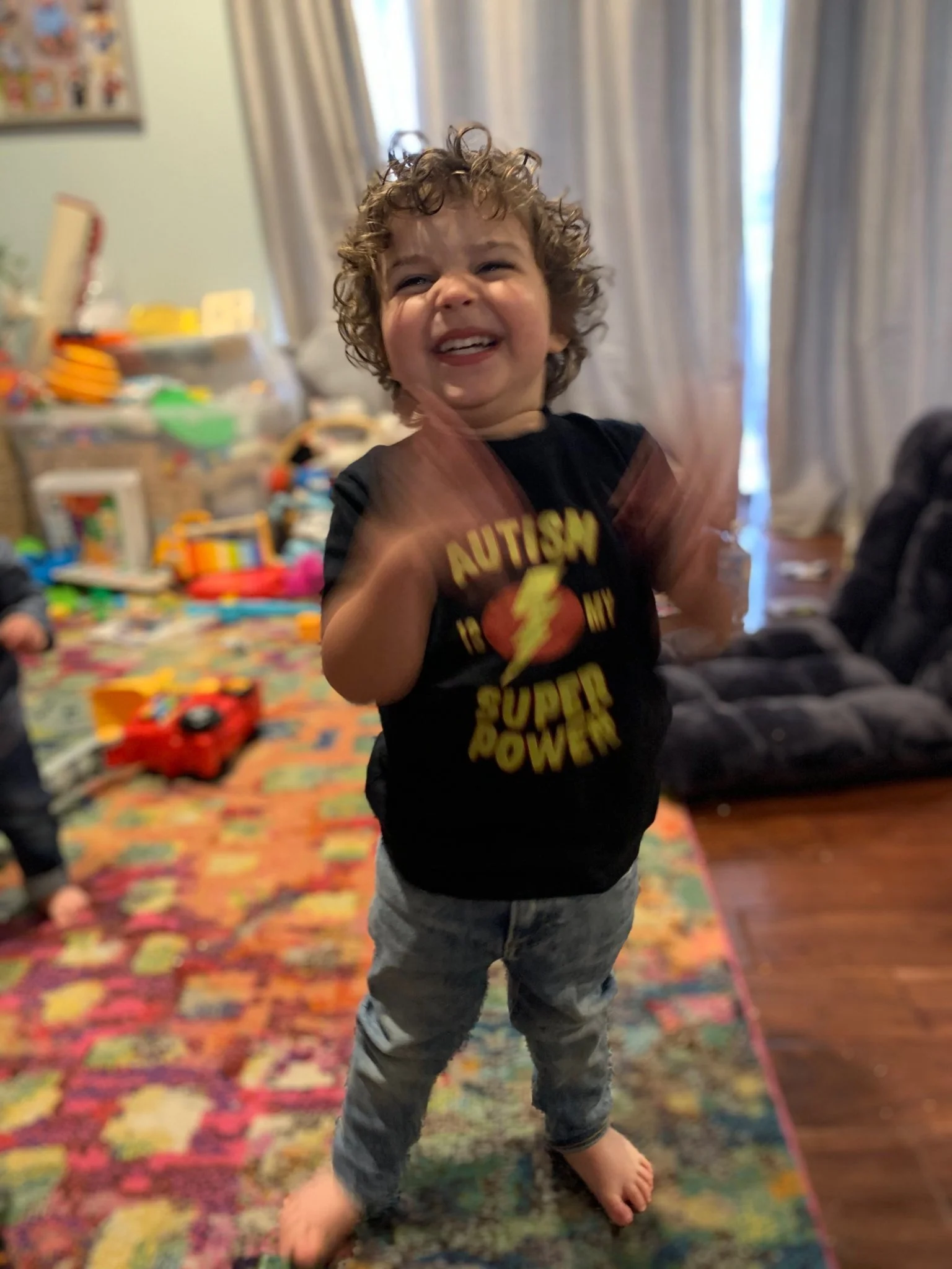 A young boy with curly hair smiling and clapping in a colorful playroom with toys and a patterned rug.