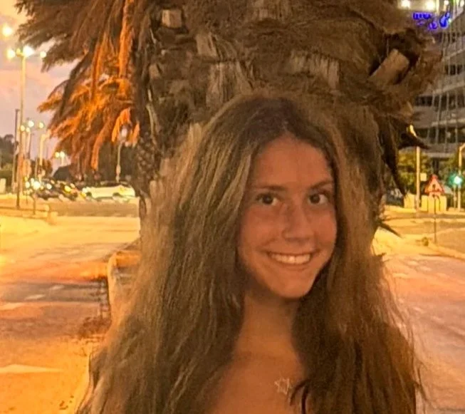 A smiling woman with long, wavy hair standing outdoors at night in front of a tree with large, textured leaves. City lights and buildings are visible in the background.