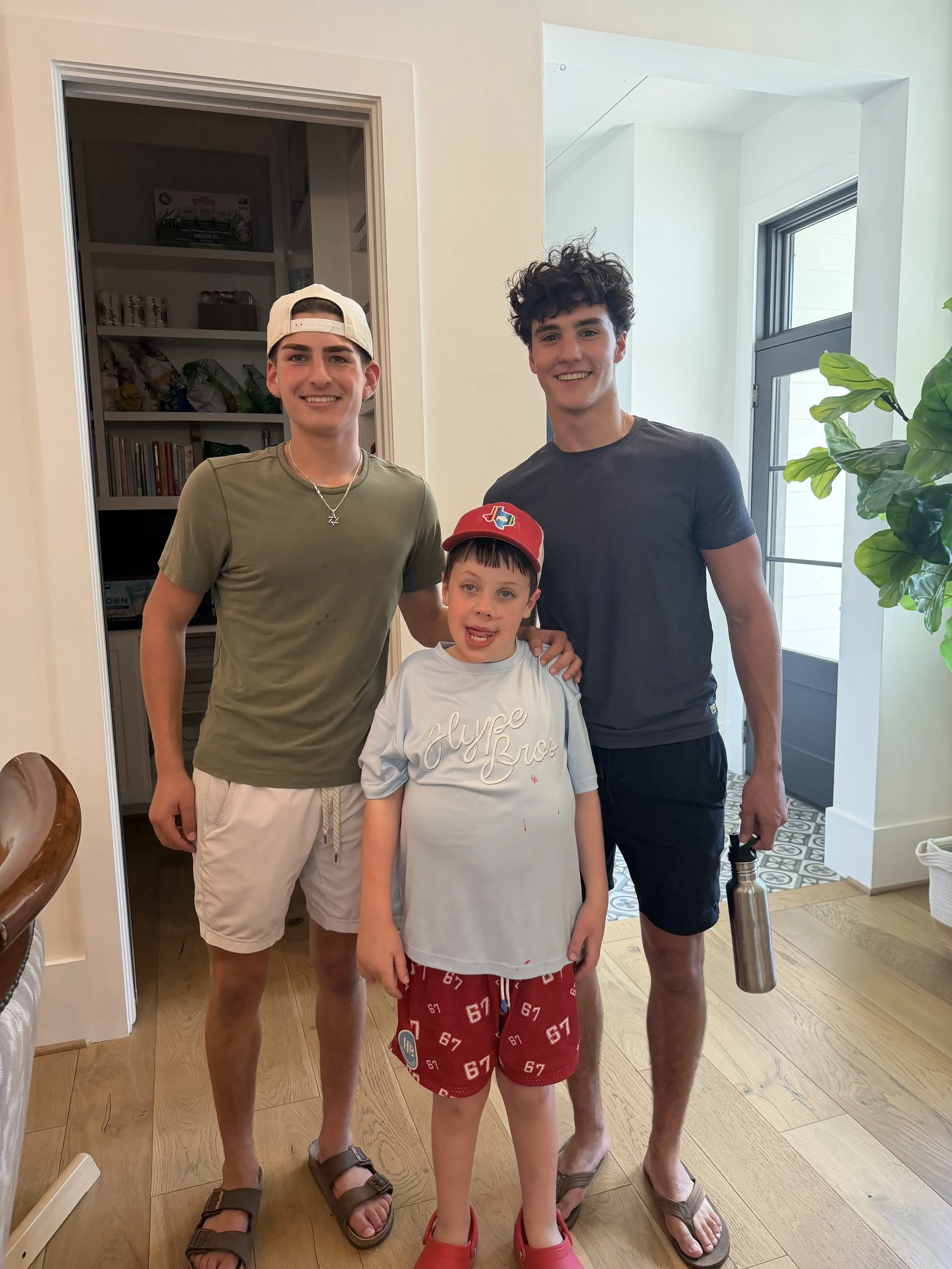 Three boys standing indoors, smiling for the camera, near a doorway and a window. The boy on the left wears a beige cap backward, a green T-shirt, beige shorts, and brown sandals. The boy in the middle is younger, wearing a white T-shirt with writing, red shorts with numbers, and a red cap with a Texas-shaped patch. The boy on the right has curly dark hair, wears a dark T-shirt, dark shorts, and is holding a water bottle. The background includes a closet and a greenery plant.