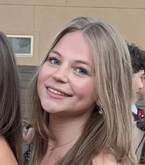 A young woman with blonde hair and blue eyes smiling at the camera, wearing a dress and earrings, at an indoor event.