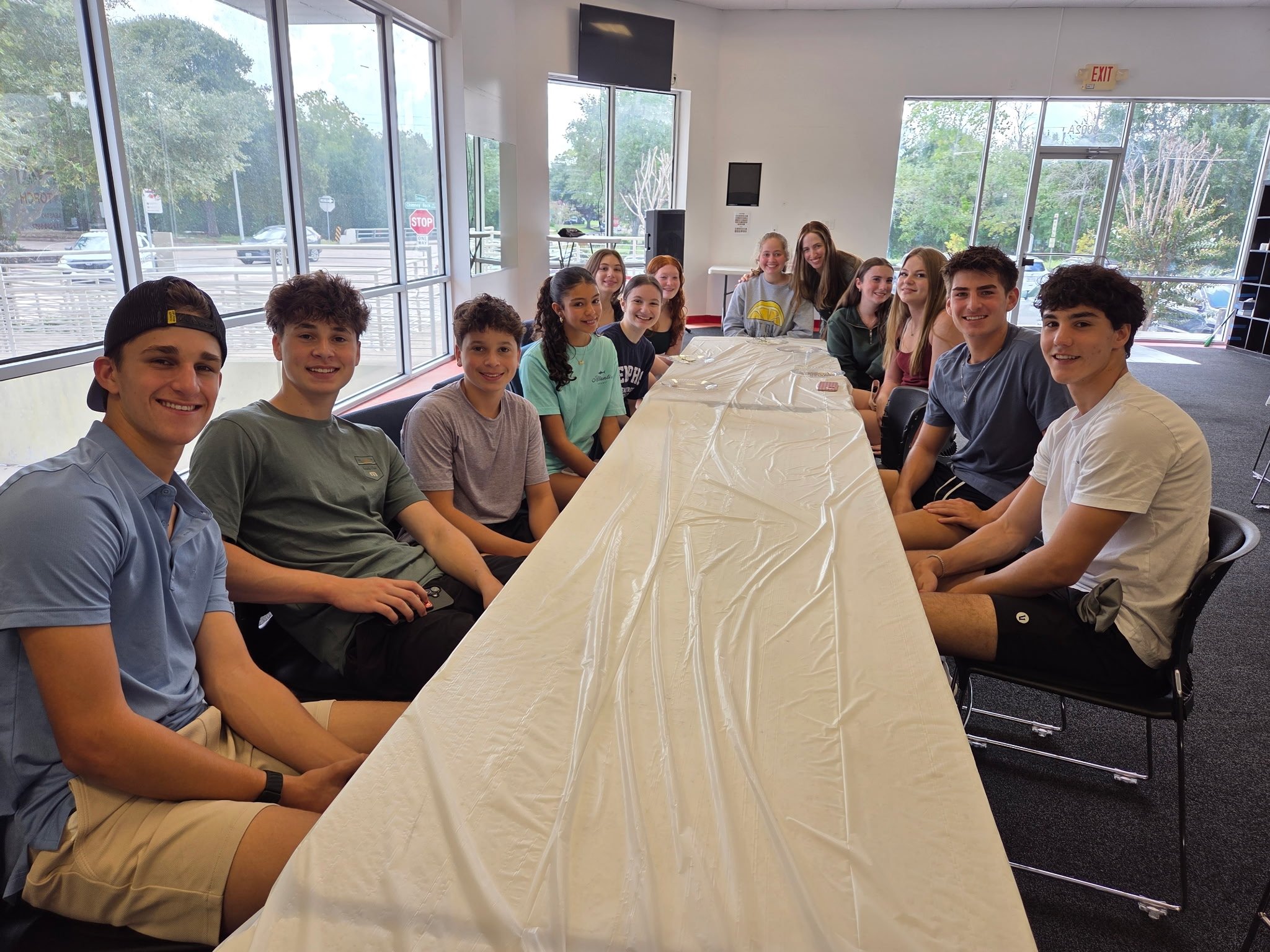 A group of young people, mostly teenagers, are sitting around a long table covered with a plastic tablecloth in a bright room with large windows. They are smiling and looking at the camera.