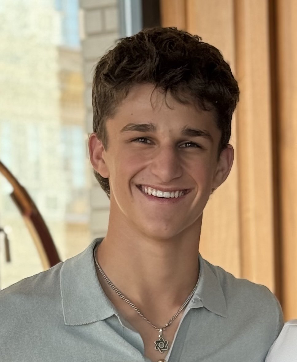 A young man smiling, wearing a light grey polo shirt, a necklace with a pendant, with a background of a window and curtain.