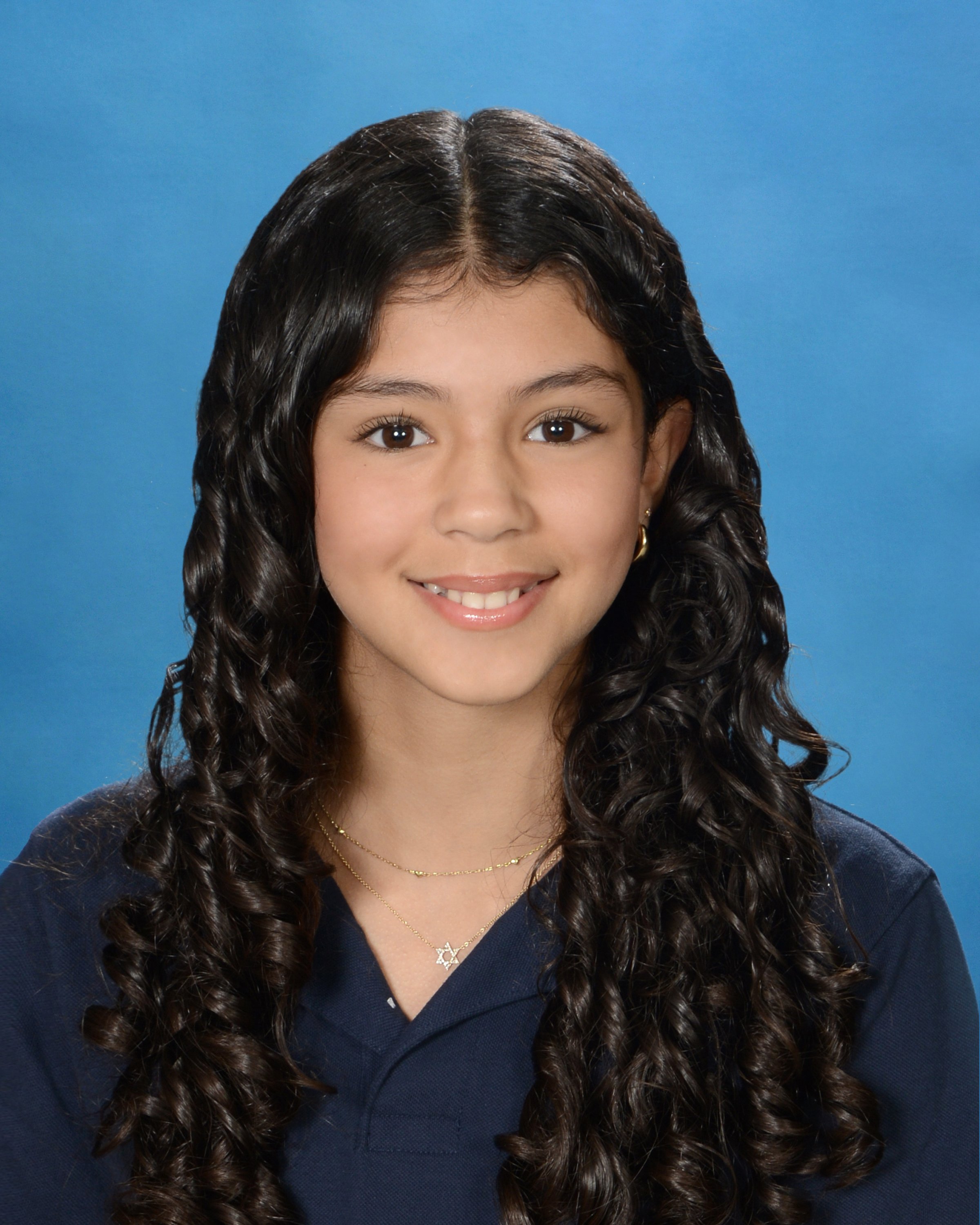 Close-up of a young woman with long, curly dark hair, brown eyes, wearing a black top and gold jewelry, against a blue background.