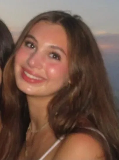 A young woman with long, wavy brown hair smiling at the camera, wearing a white top, during sunset.