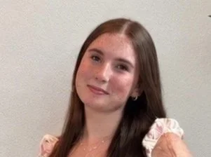 Young woman with long brown hair wearing a light-colored blouse, smiling gently at the camera.